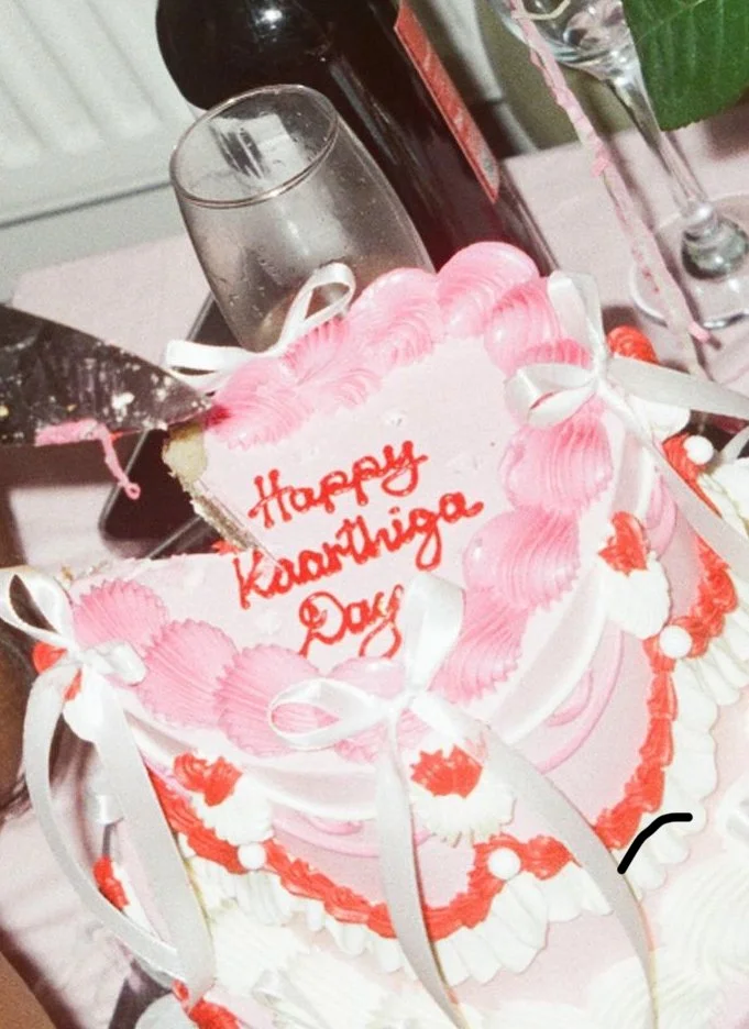 A birthday cake decorated with pink and white frosting, with a message written in red icing that says 'Happy Kaarthiga Day.' The cake is adorned with white ribbons and red decorative accents. There are glasses and a bottle in the background.
