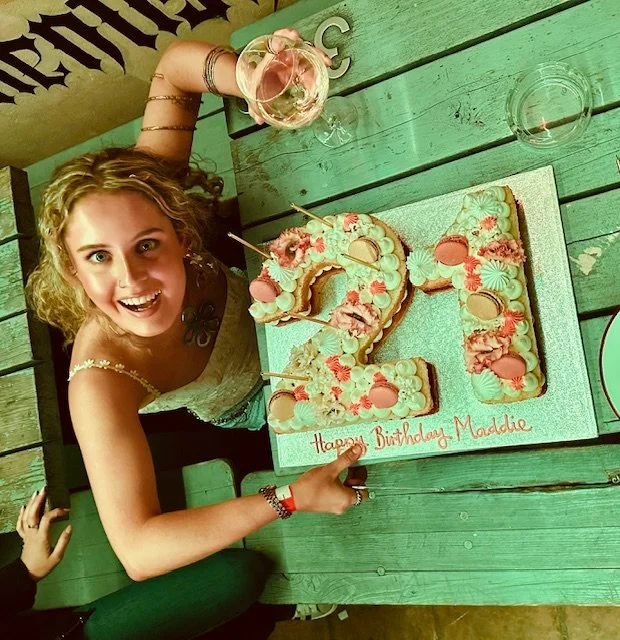 Young girl with curly hair smiling at a birthday celebration, sitting at a green wooden table with a cake shaped as the number 13, decorated with pink and white frosting and flowers. There is a glass of wine next to her on the table.