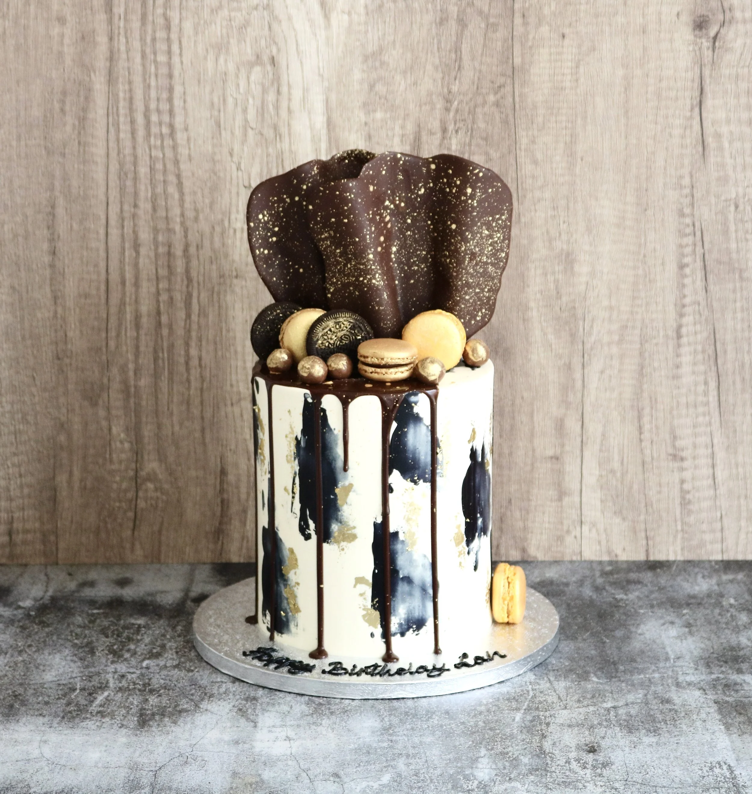 Luxury black and gold birthday cake with chocolate drip, edible gold leaf, gold-painted macarons, handmade chocolate toppers, and artistic buttercream finish for celebrations in London.