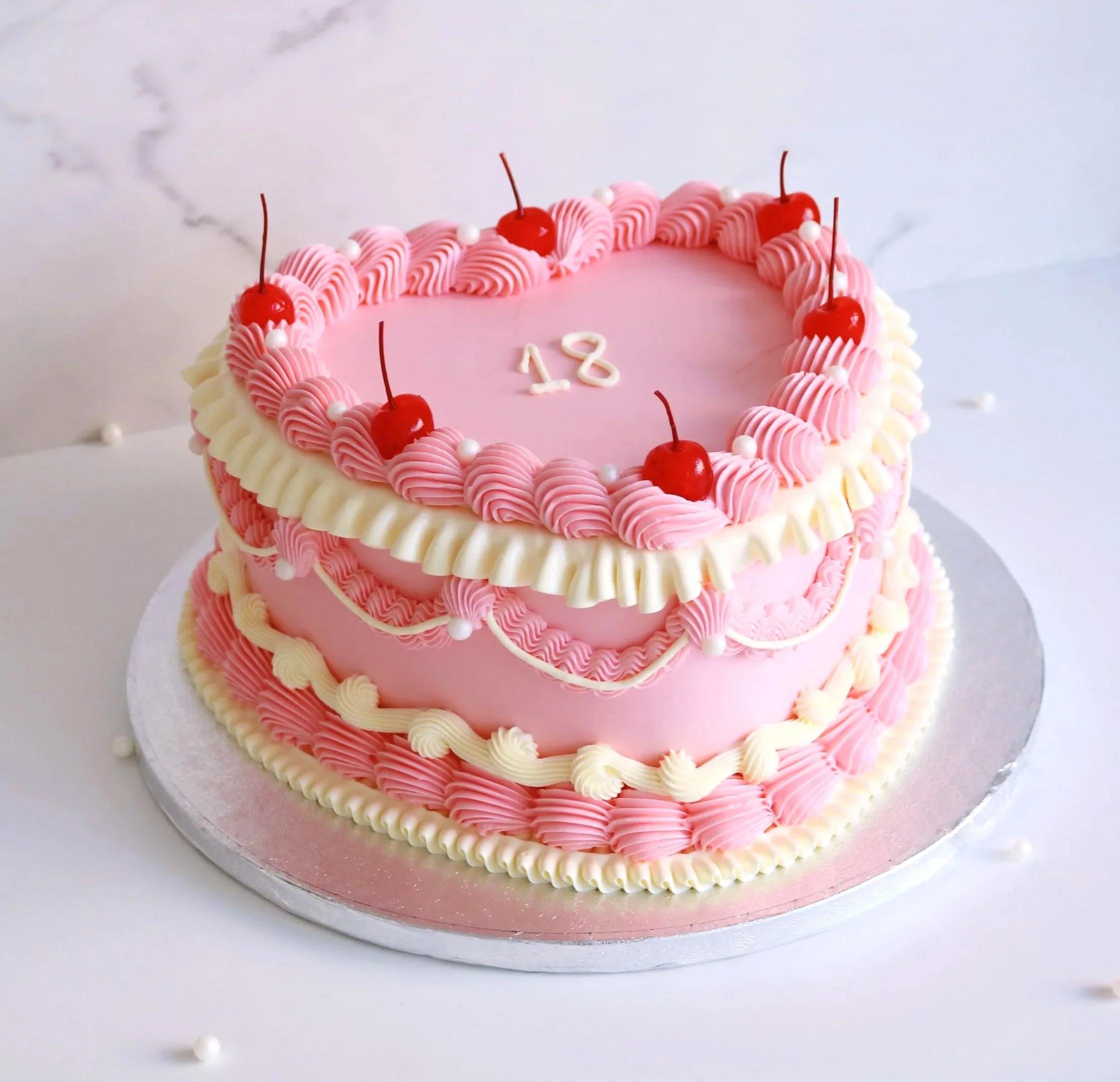 Heart-shaped pink birthday cake with pink and white icing, decorated with cherries on top and red icing accents, with the message 'Happy 17th Grace & Tash' written in red icing.