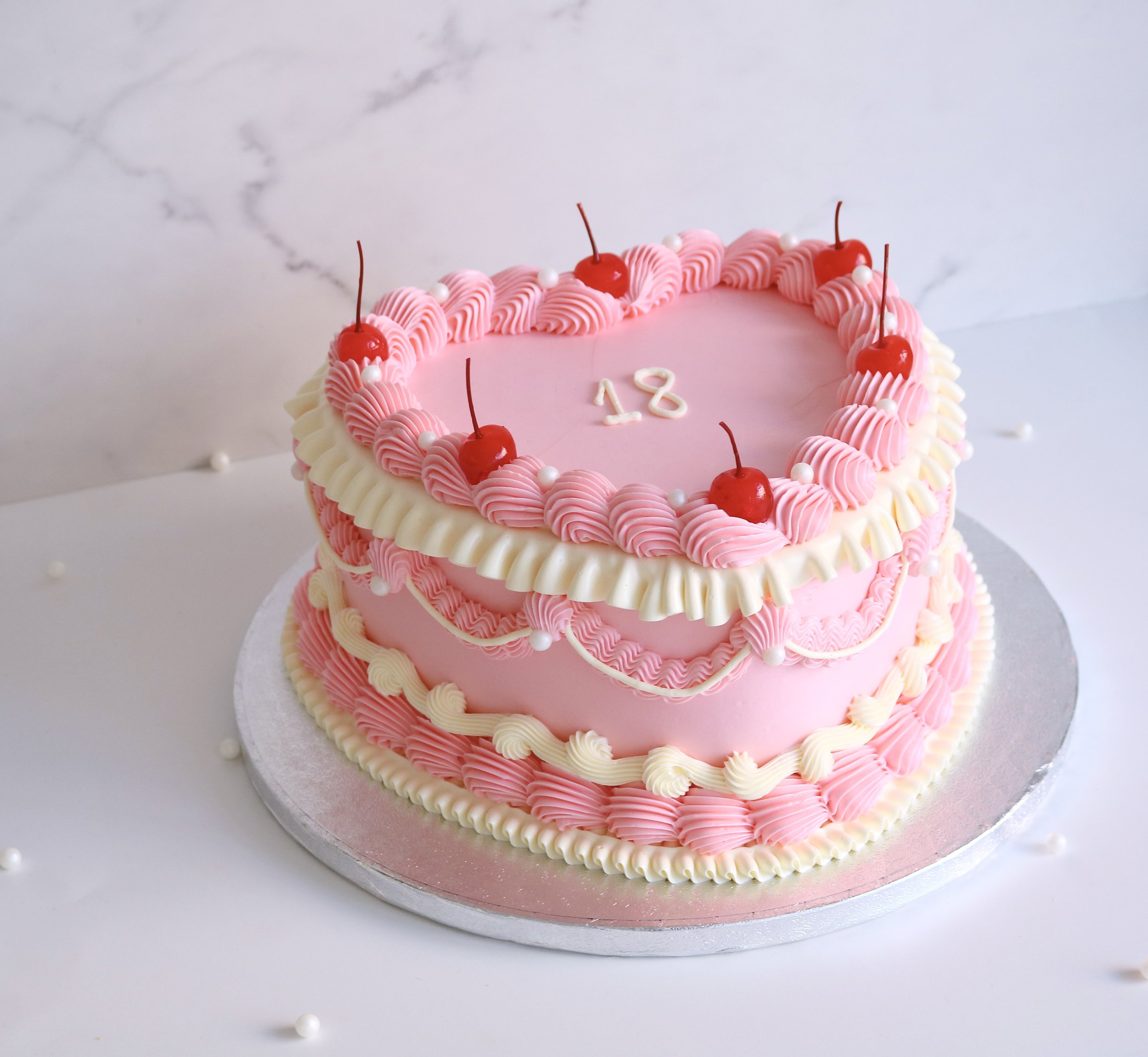 vintage cake in heart shape, decorated with intricate ruffles in light pink and cream colour scheme, decorated with red cherries and pearls around.