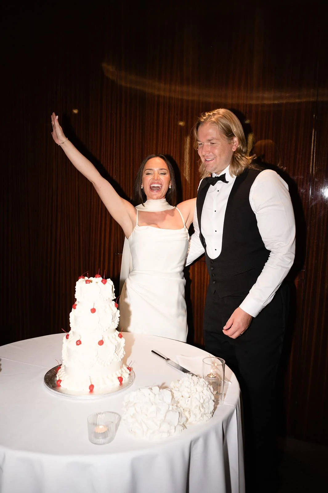 A bride and groom celebrating their wedding with a tiered white cake topped with cherries.