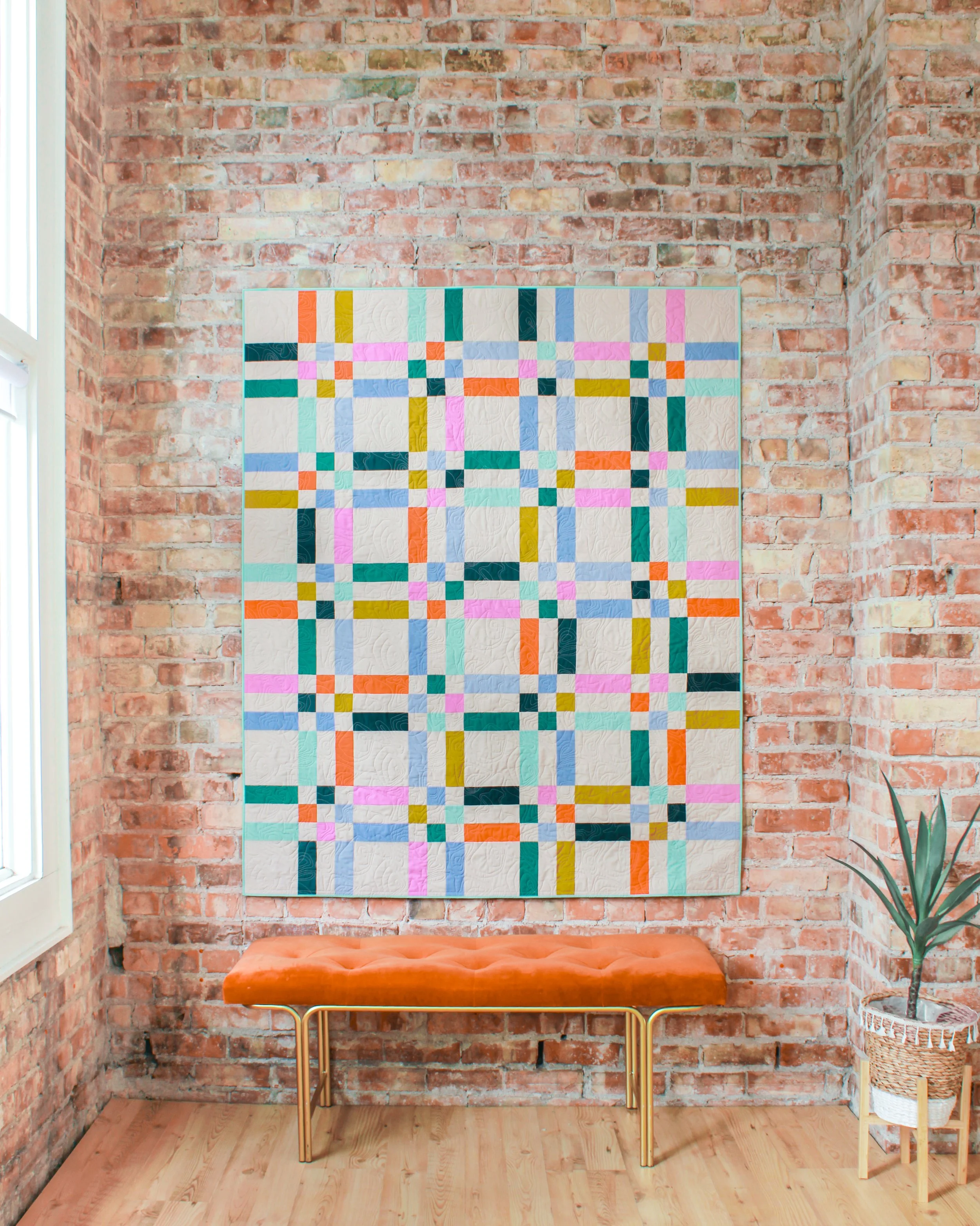 A plaid-like design quilt hanging on a white brick wall with a geometric pattern in shades of blue, white, pink, brown, and tan.