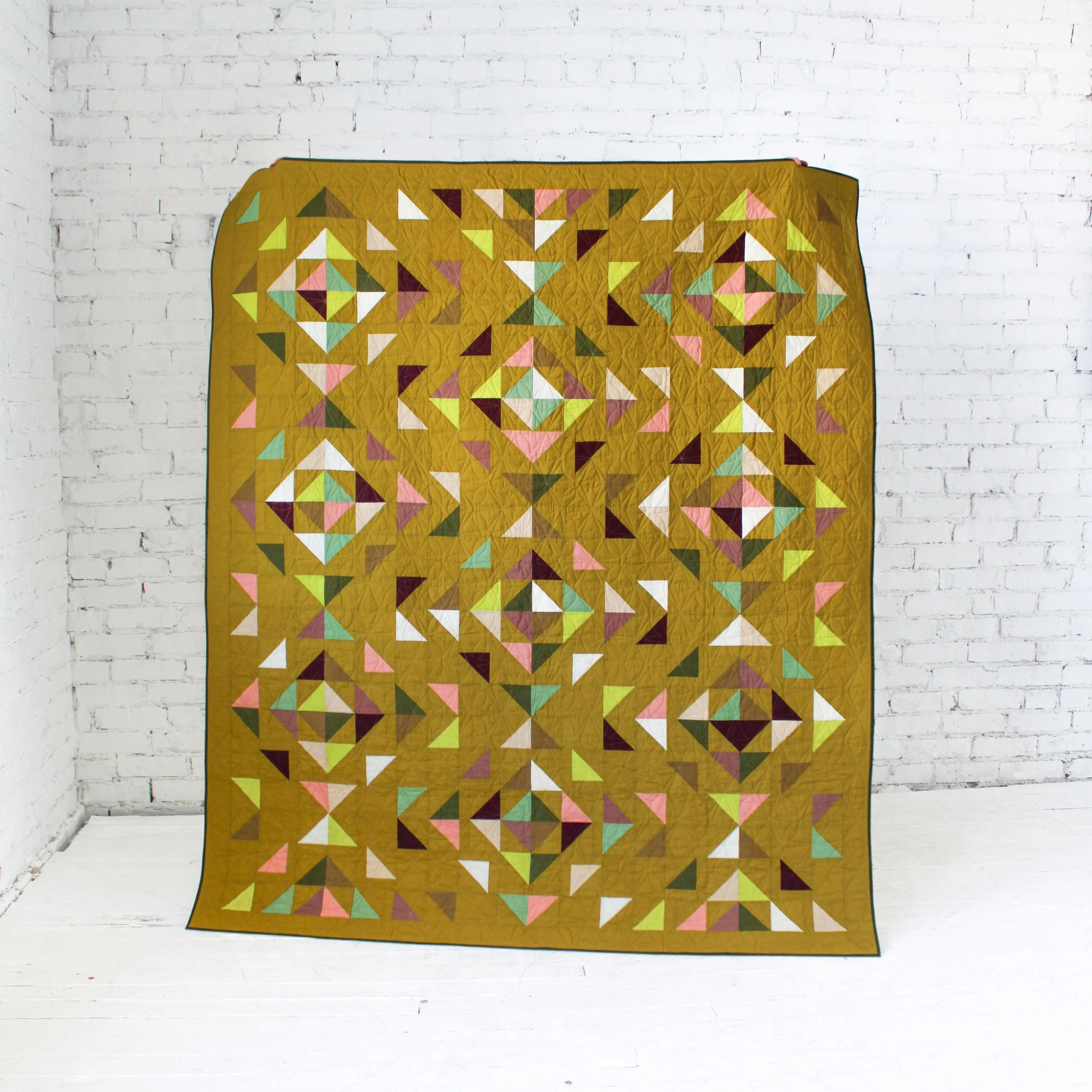 A colorful quilt with a pattern of triangles in shades of pink, yellow, green, white, and burgundy, hanging on a white brick wall.