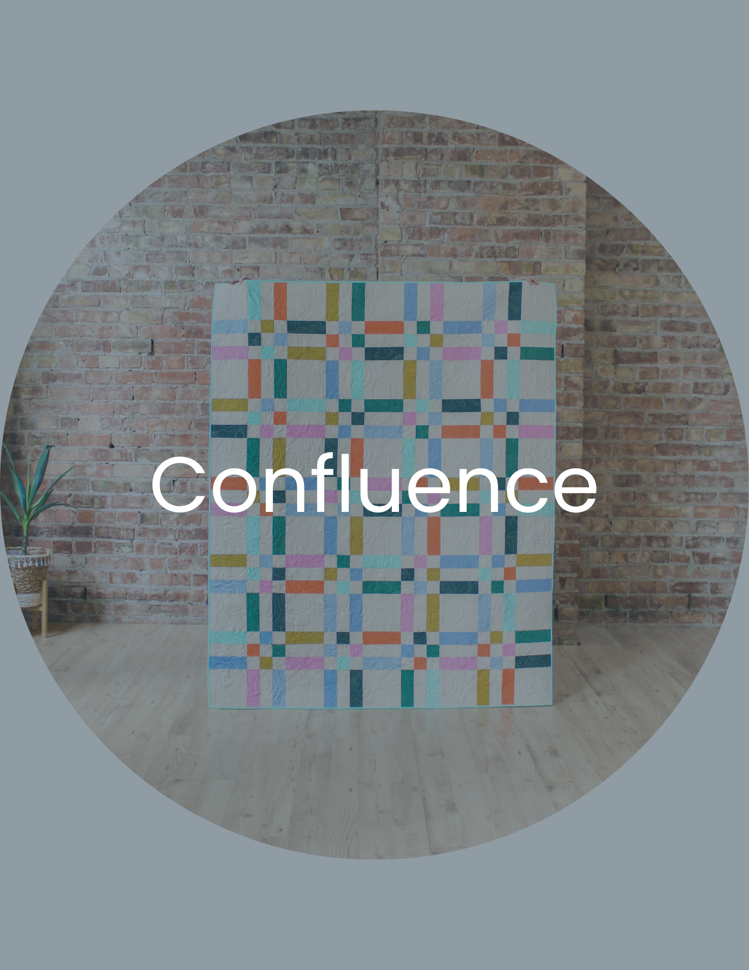 A colorful quilt featuring a geometric pattern of squares and rectangles in various pastel and vibrant colors, hanging in front of an exposed brick wall with a wooden floor and a potted plant nearby.
