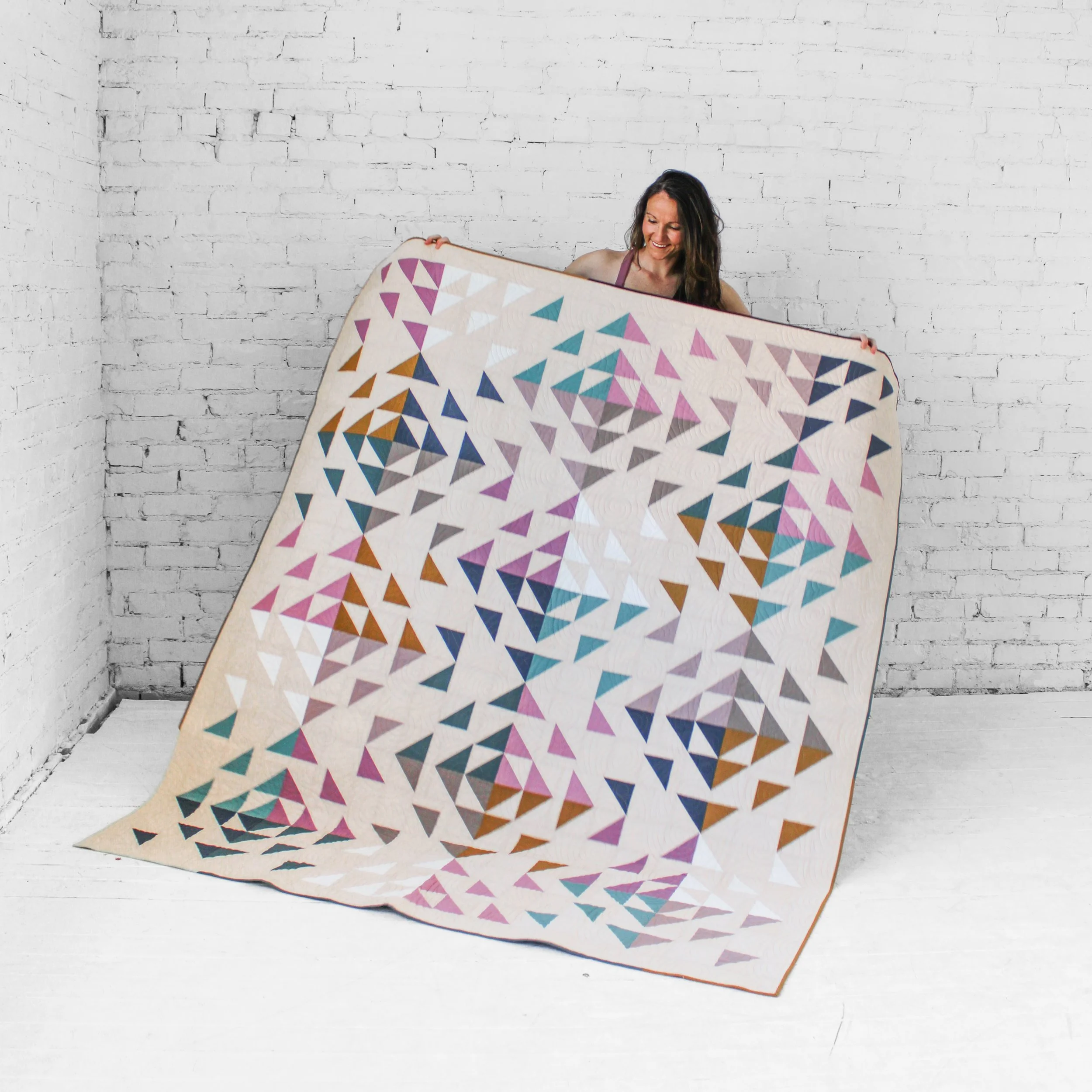 A woman is holding a colorful quilt with geometric patterns in front of a white brick wall.