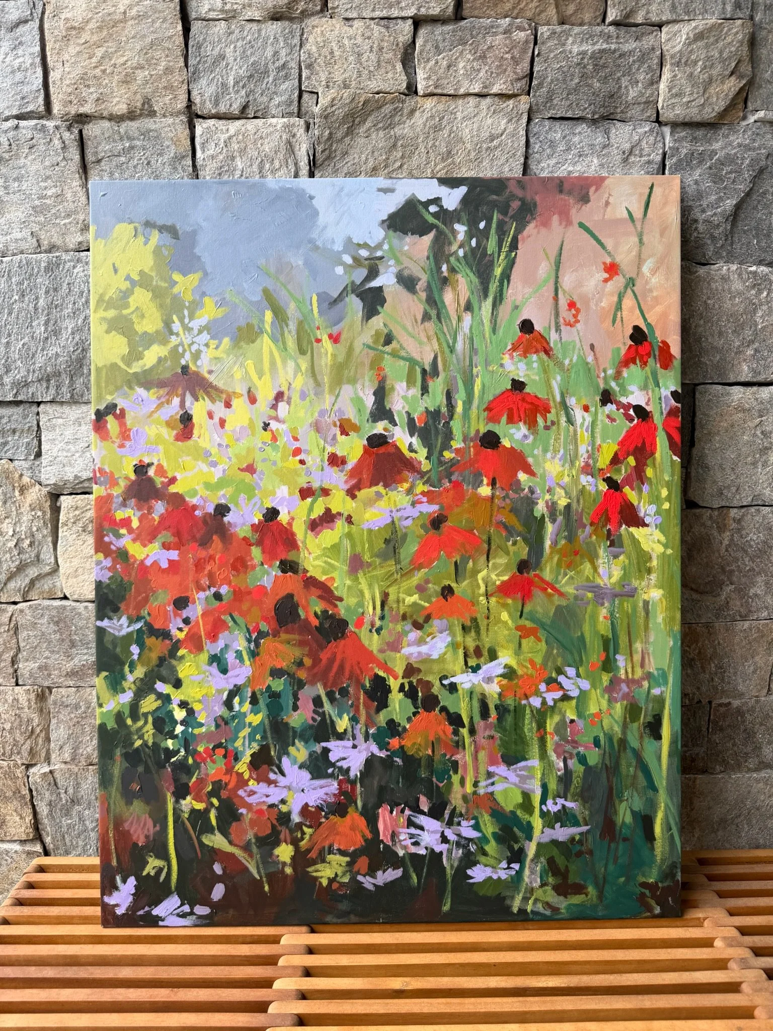 Detail of a framed mixed media floral painting by Cheyenne Trunnell, showing red-orange flowers, tall stems, and soft peach and green layers against a stone wall.