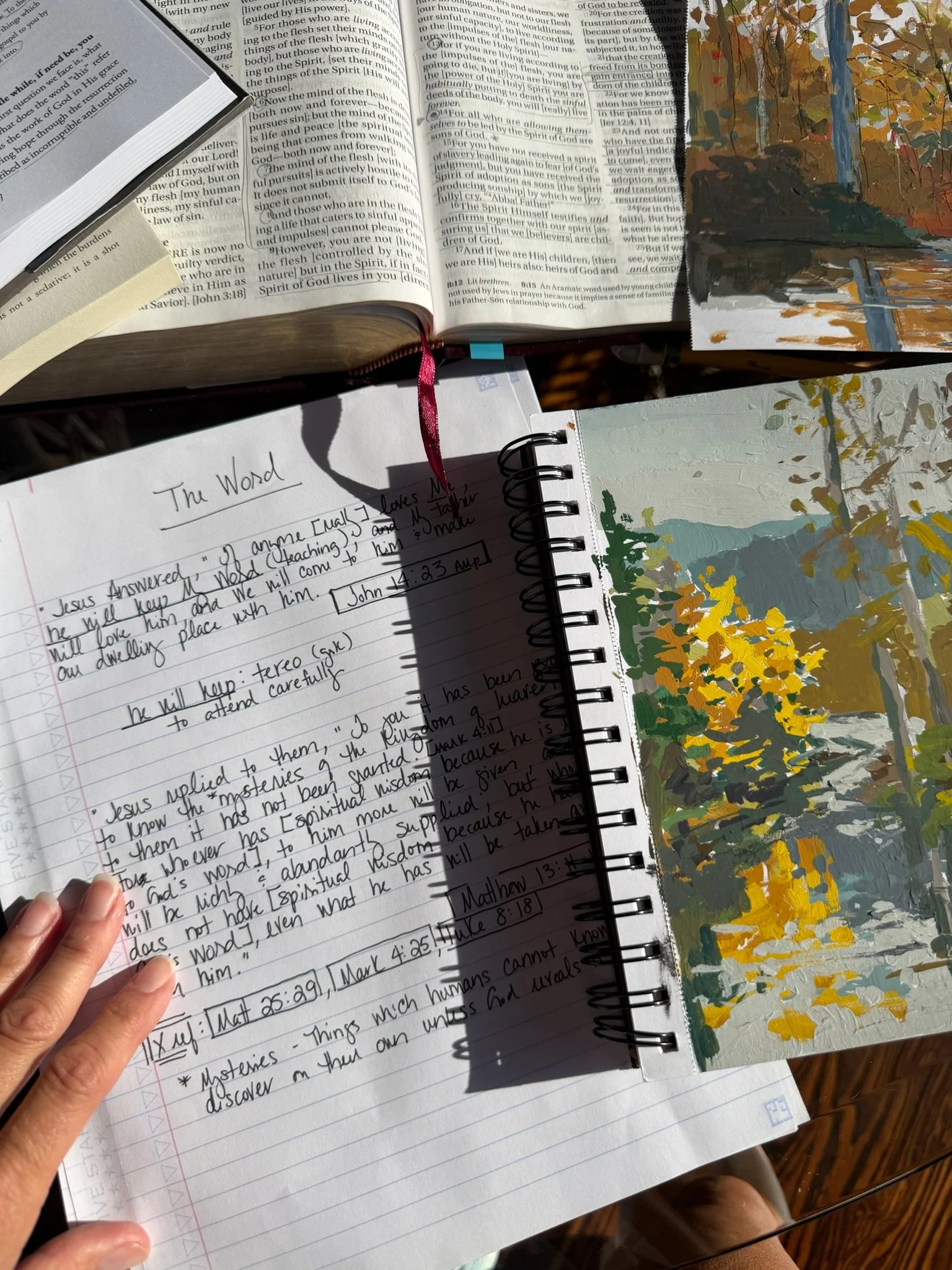 Open Bible and handwritten study notes titled “The Word” beside painted landscape studies in warm sunlight.