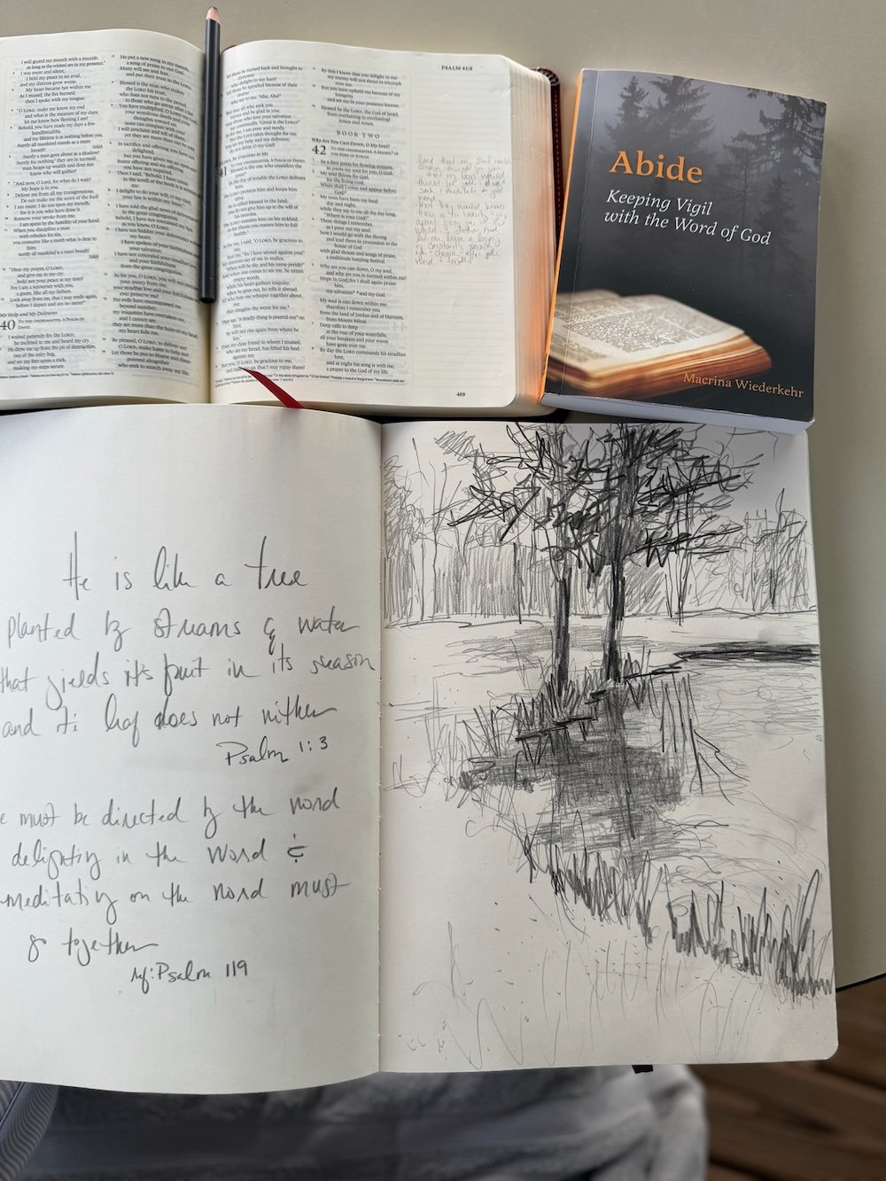 Open sketchbook with charcoal tree sketches and handwritten Scripture, photographed alongside an open Bible and devotional book during a winter studio journaling practice.