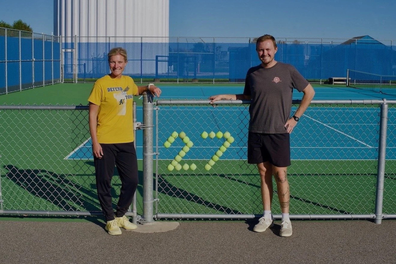 Two Hastings tennis coaches stand beside a court gate decorated with tennis balls.
