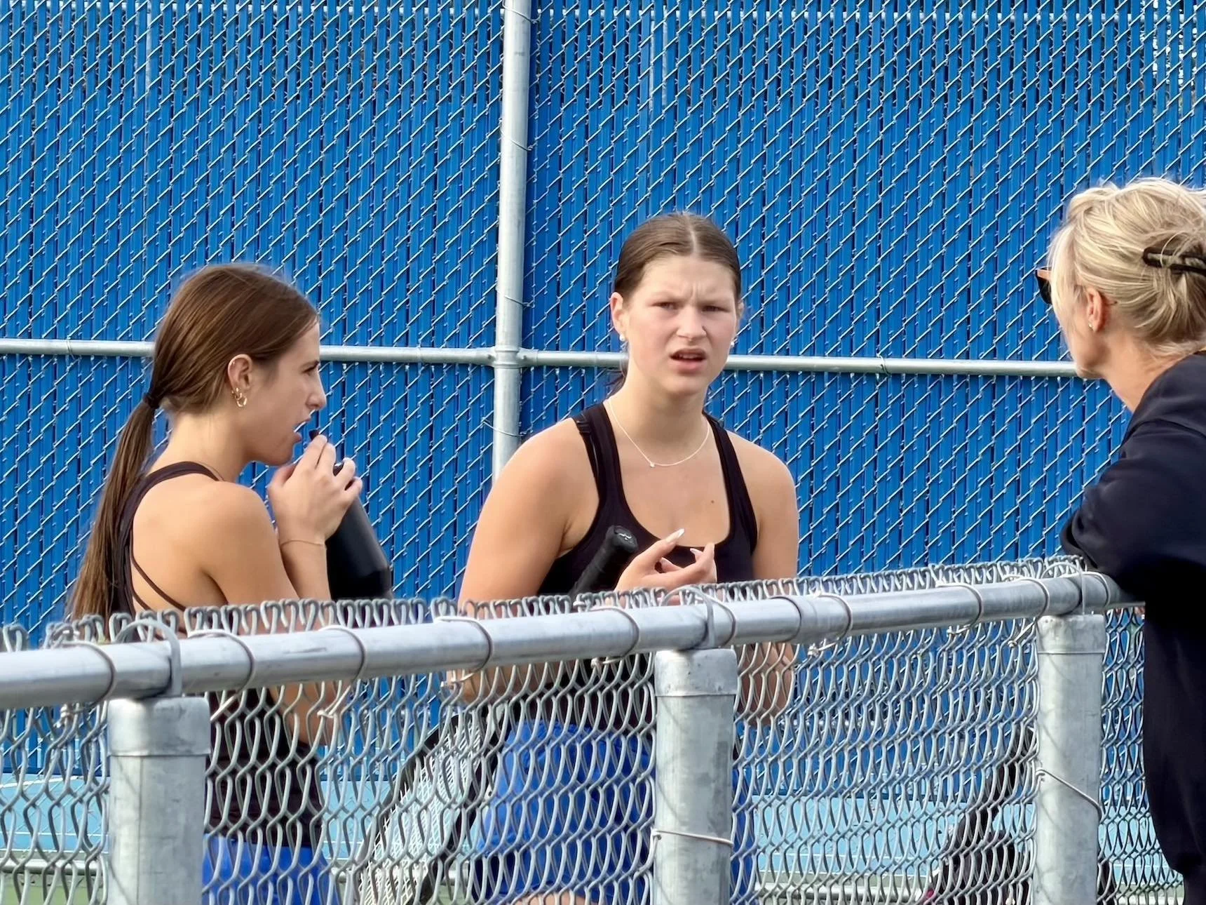 Two tennis players speak with a coach beside a chain-link court fence.