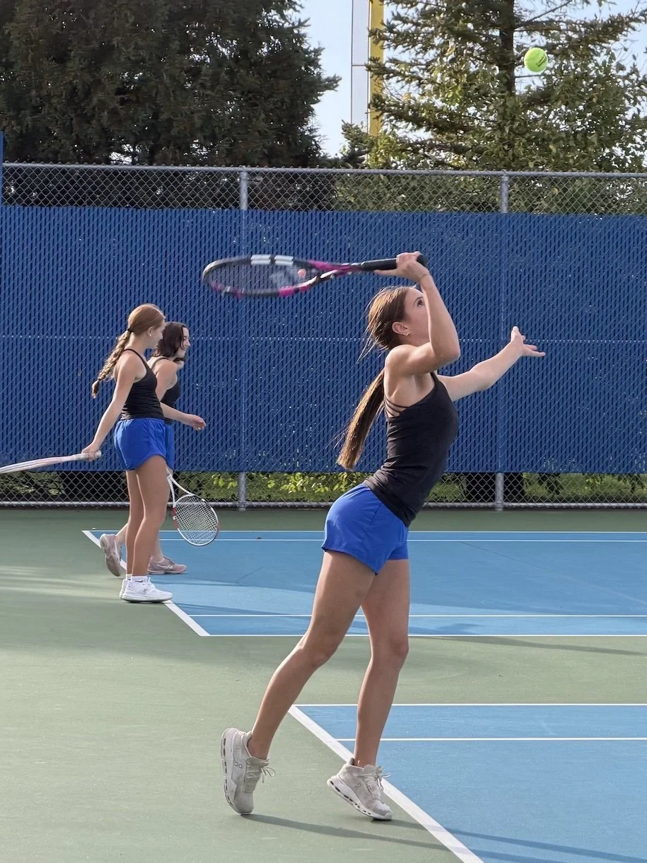 A Hastings tennis player raises her racket beneath a ball in the air while two players stand behind her.