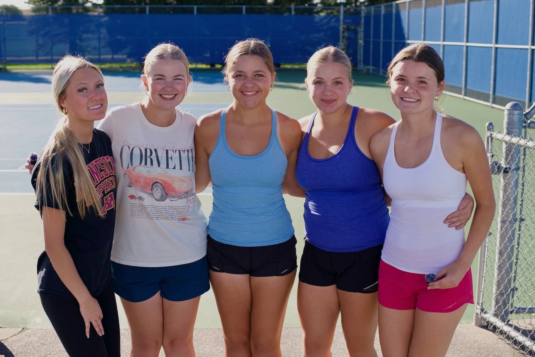 Five Hastings girls tennis players stand arm in arm beside outdoor tennis courts.