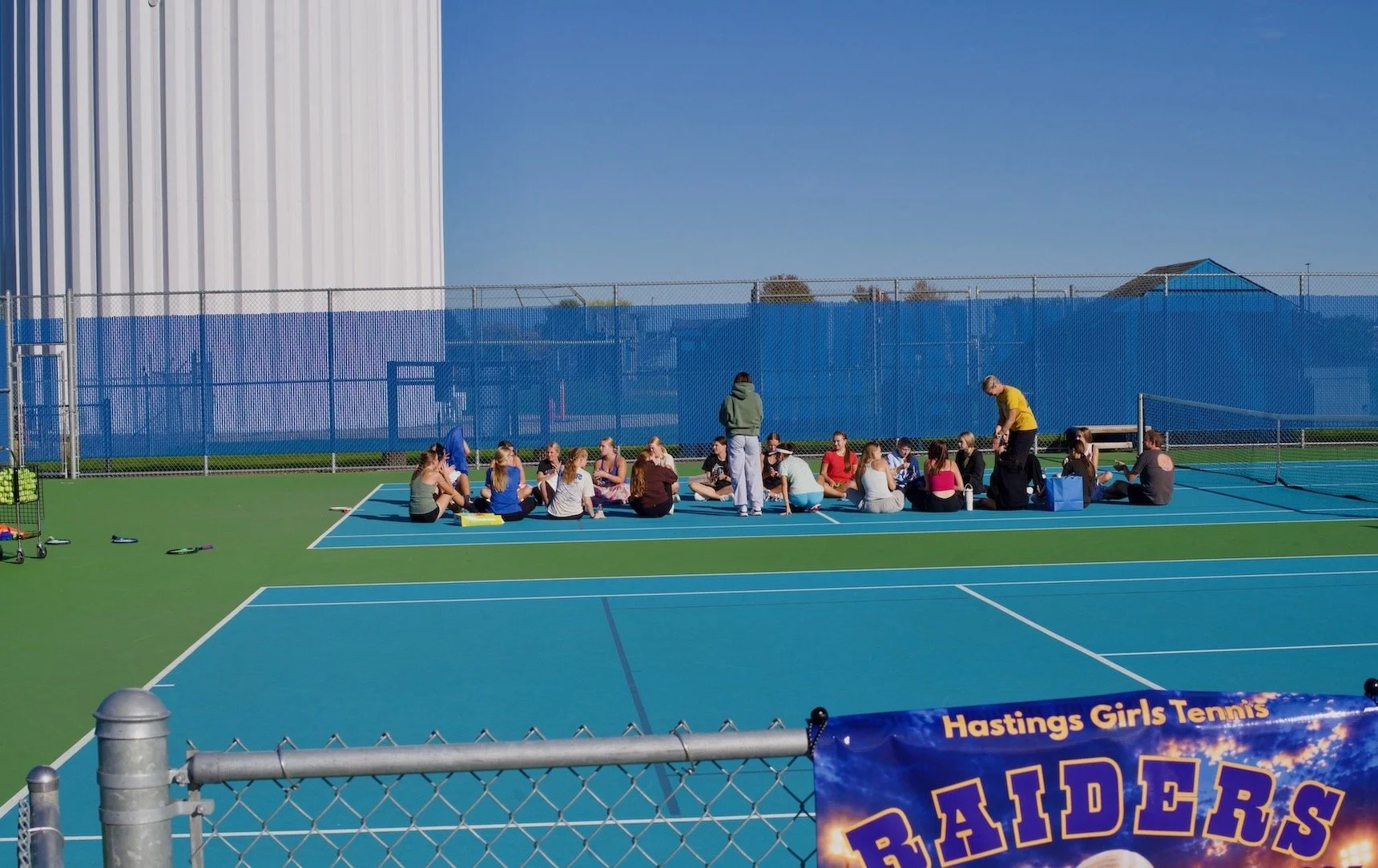Players sit in a circle on a blue tennis court with coaches nearby.