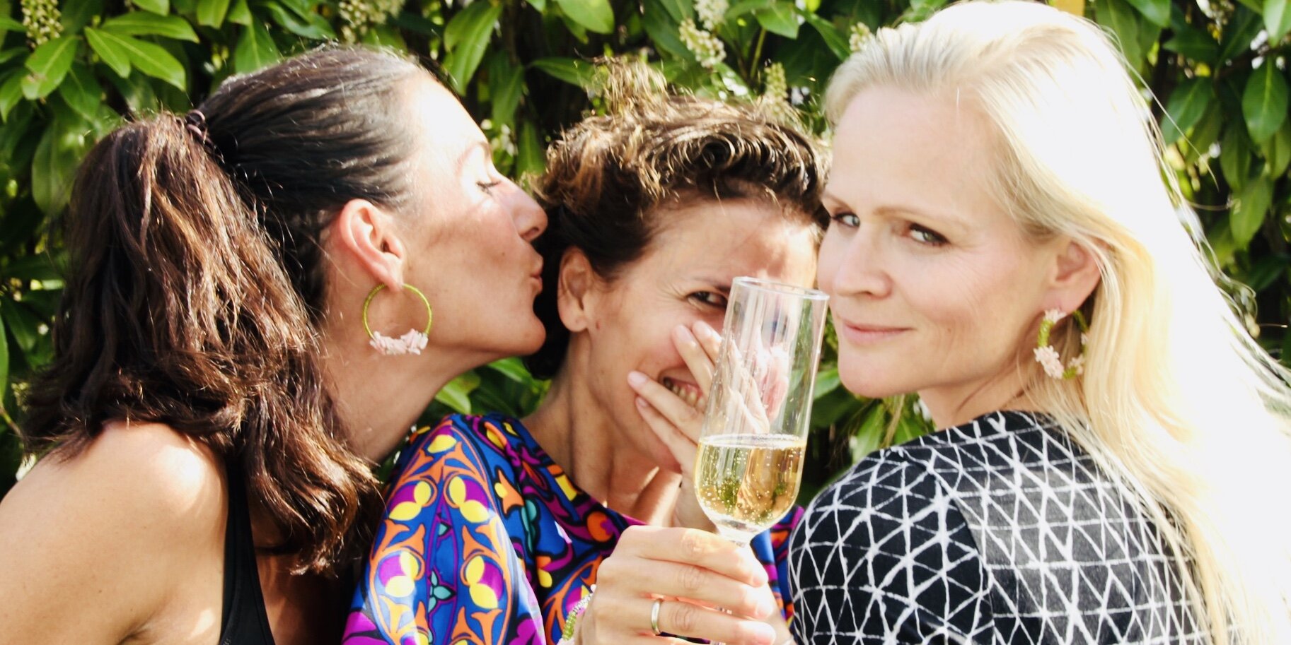 Drei Frauen, die draußen lachen und plaudern, während eine ein Glas Sekt hält; die Umgebung ist grün und sommerlich. Das ist das Frohlotte-Lebensgefühl mit bunten, upgecycleten Schmuckstücken von Frohlotte.
