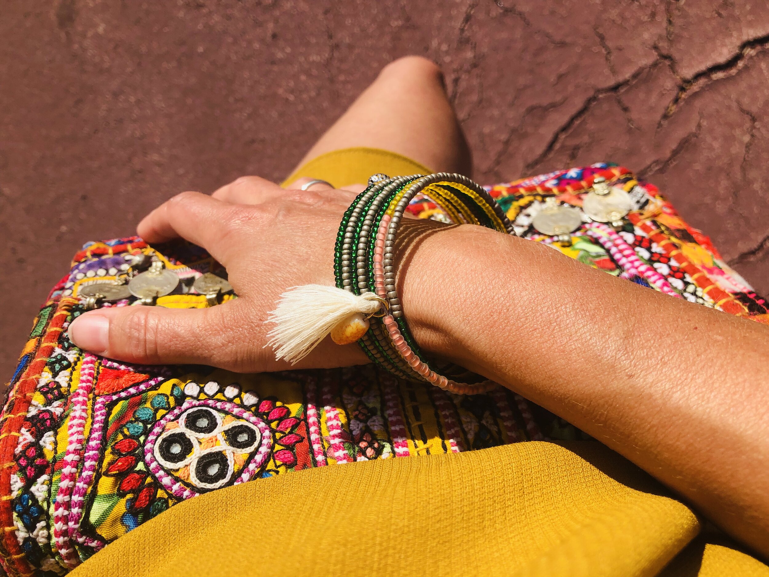 Hand mit handgemachtem, upgecycletem Armreif von Frohlotte, hält eine bunte, bestickte Tasche. Eine Person trägt ein gelbes Kleid. Der Hintergrund ist aus braunem, geplatztem Boden.