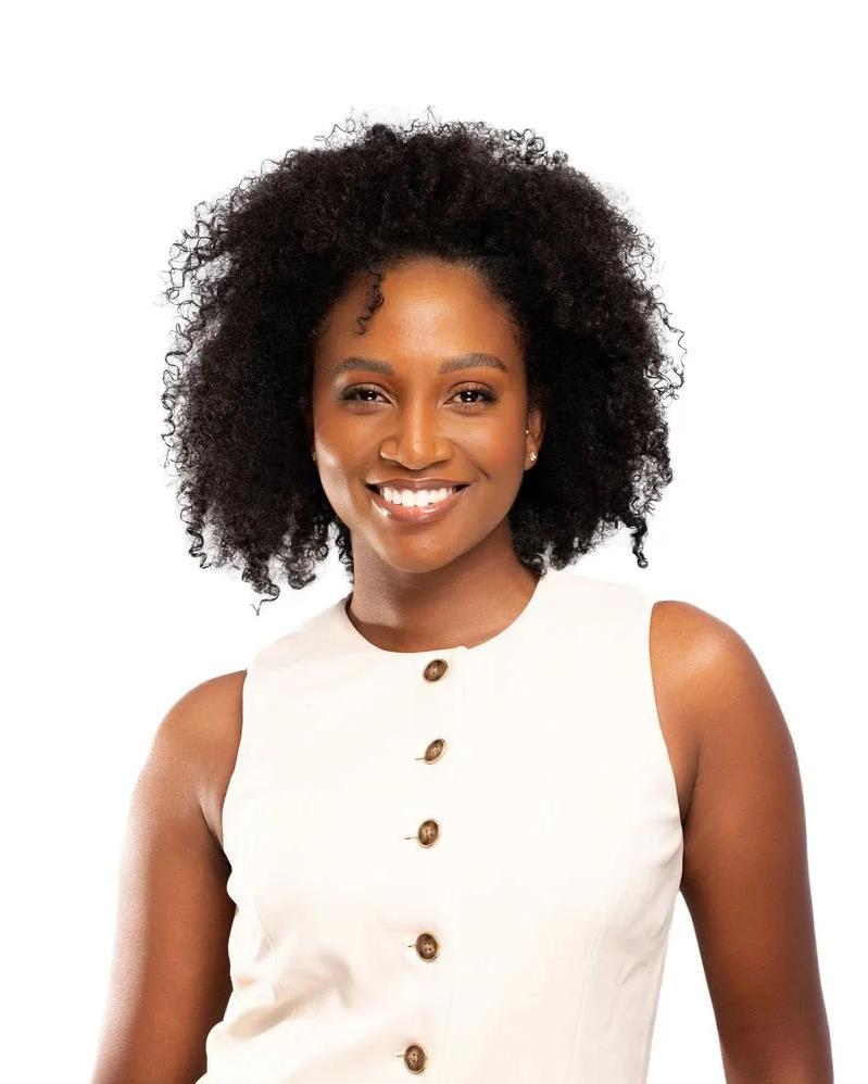 Portrait of a smiling woman with curly black hair, wearing a sleeveless white button-up top, against a plain white background.