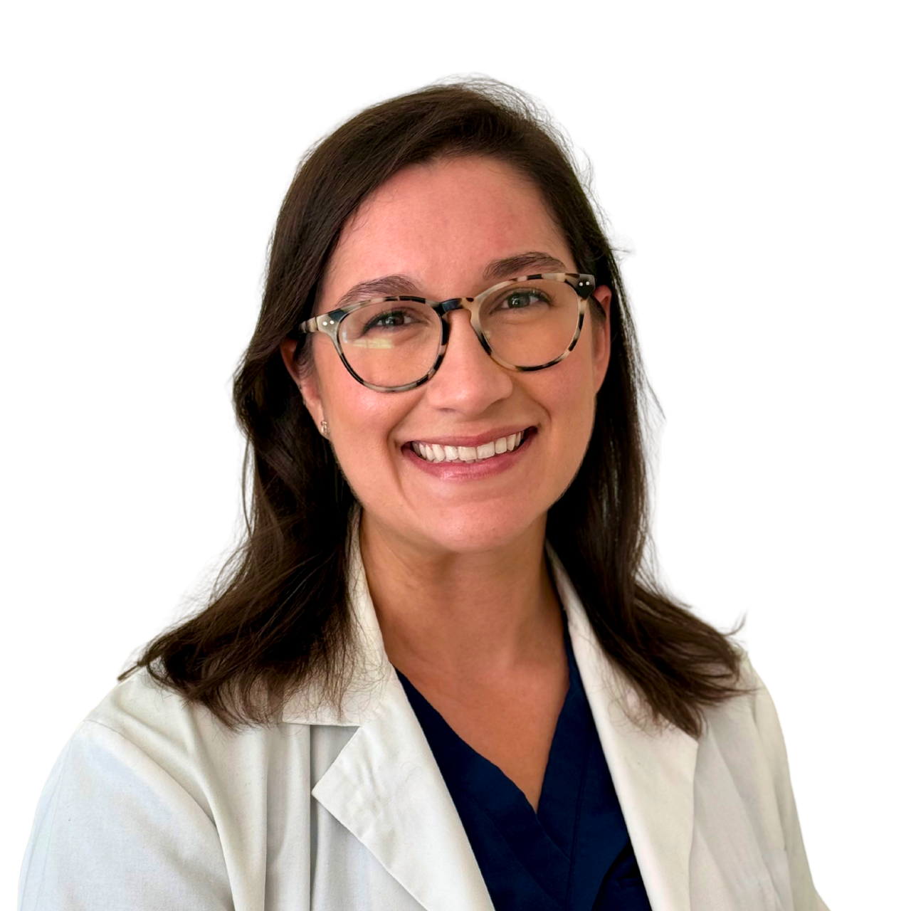 A woman with dark brown hair, glasses, and a bright smile wearing a white lab coat over dark scrubs inside a medical or clinical setting.
