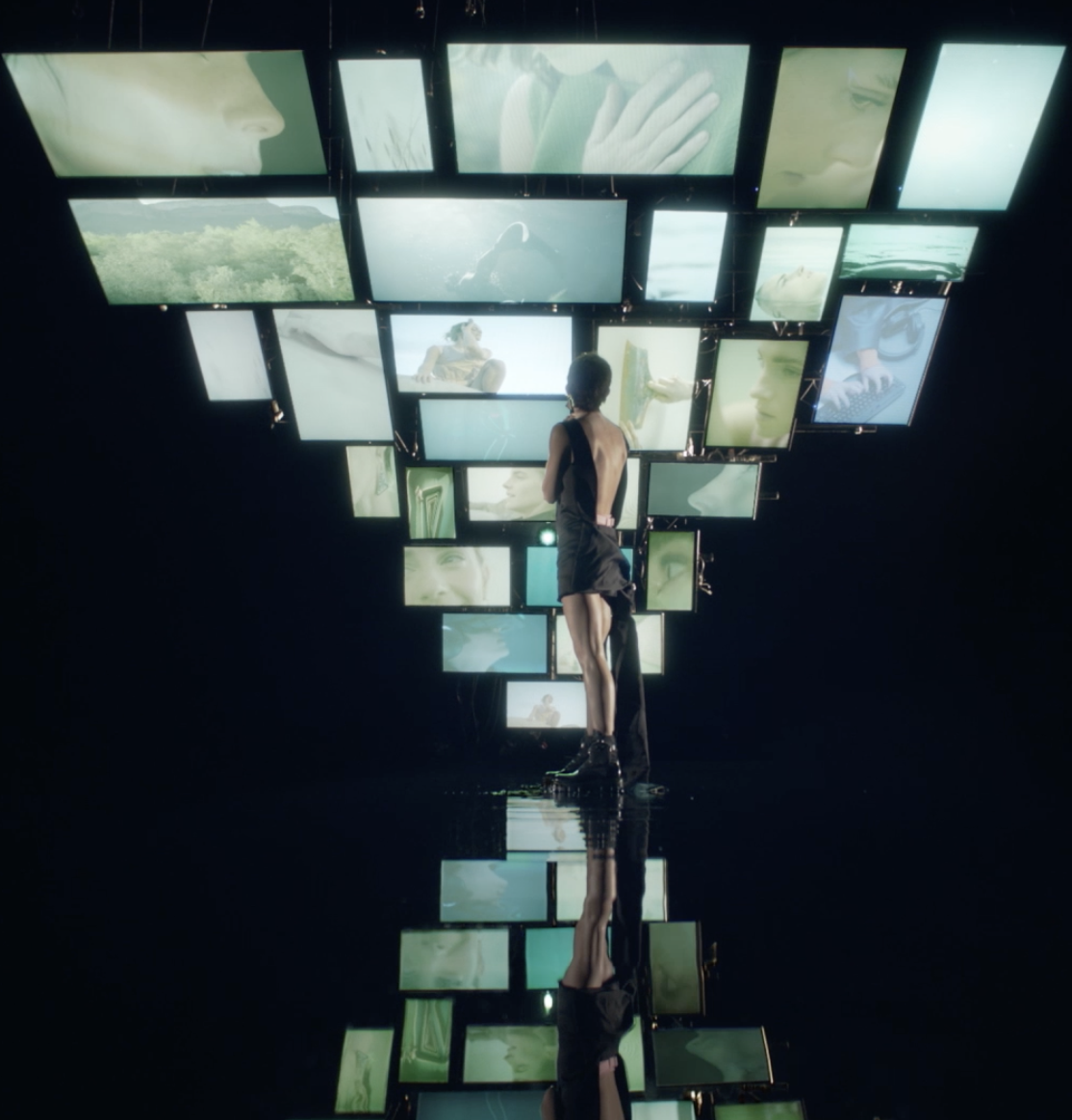 Une personne debout devant un mur de plusieurs écrans affichant des images floues ou artistiques, avec leur reflet sur un sol noir brillant.