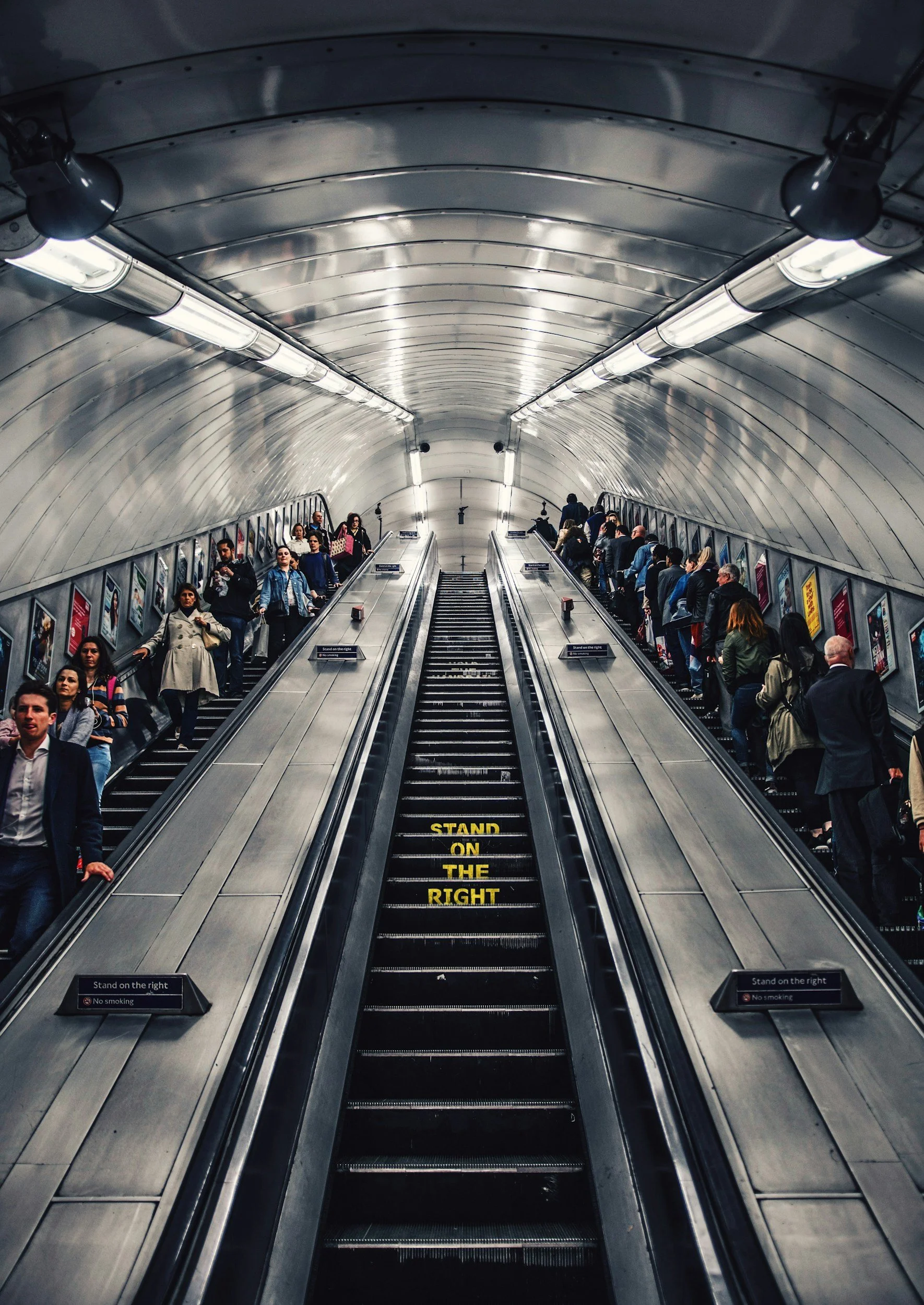 People using escalators in a subway station, with a sign on the escalator that reads 'STAND ON THE RIGHT' and people walking on the left.