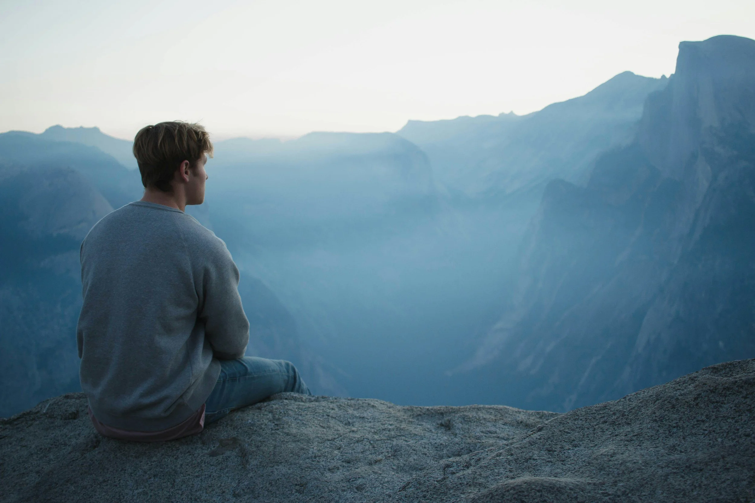 A young man sitting on a large rock, looking out over a valley with steep mountain cliffs in the distance, during dusk or dawn.