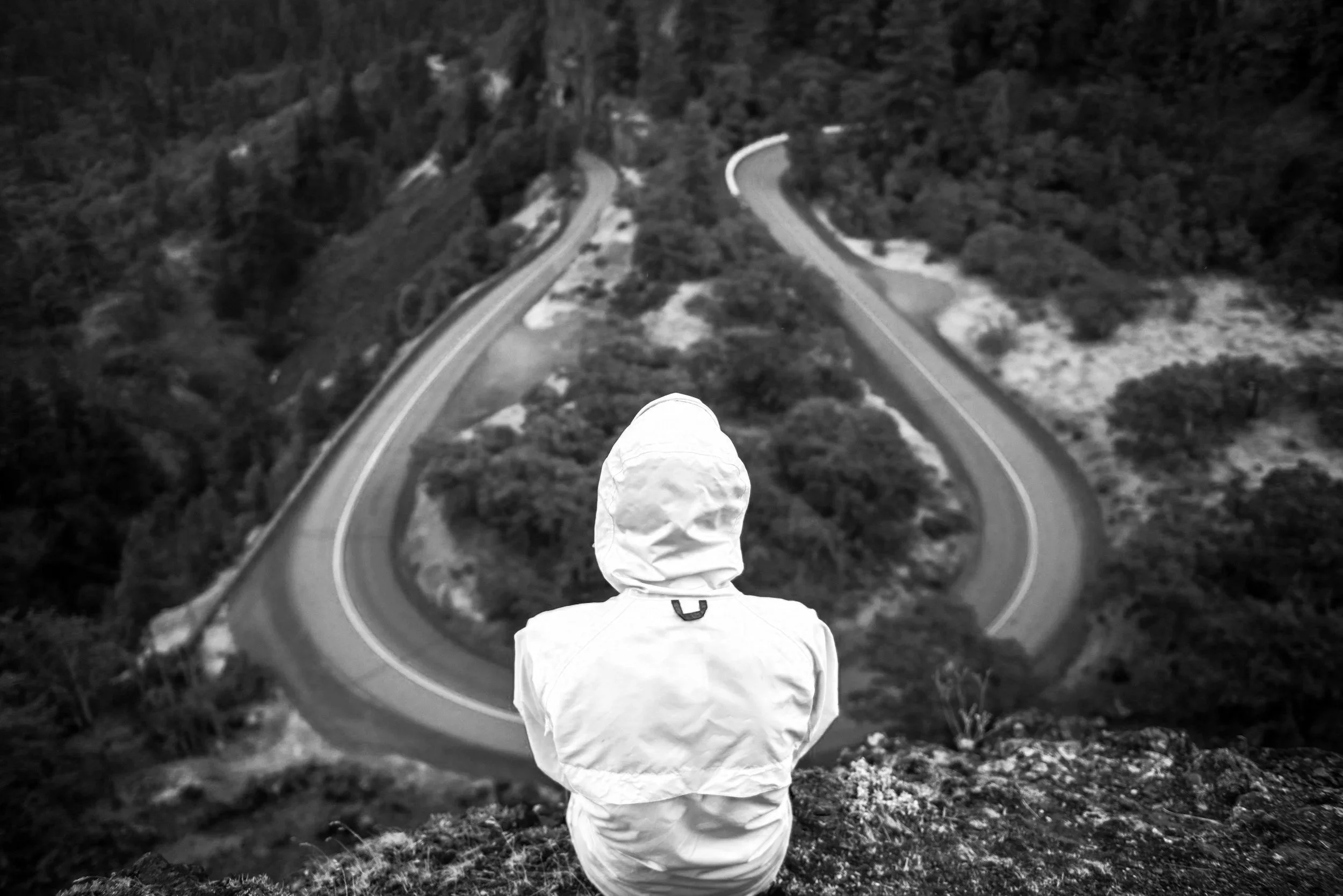 Person wearing a hooded jacket sitting on a cliff overlooking a winding mountain road