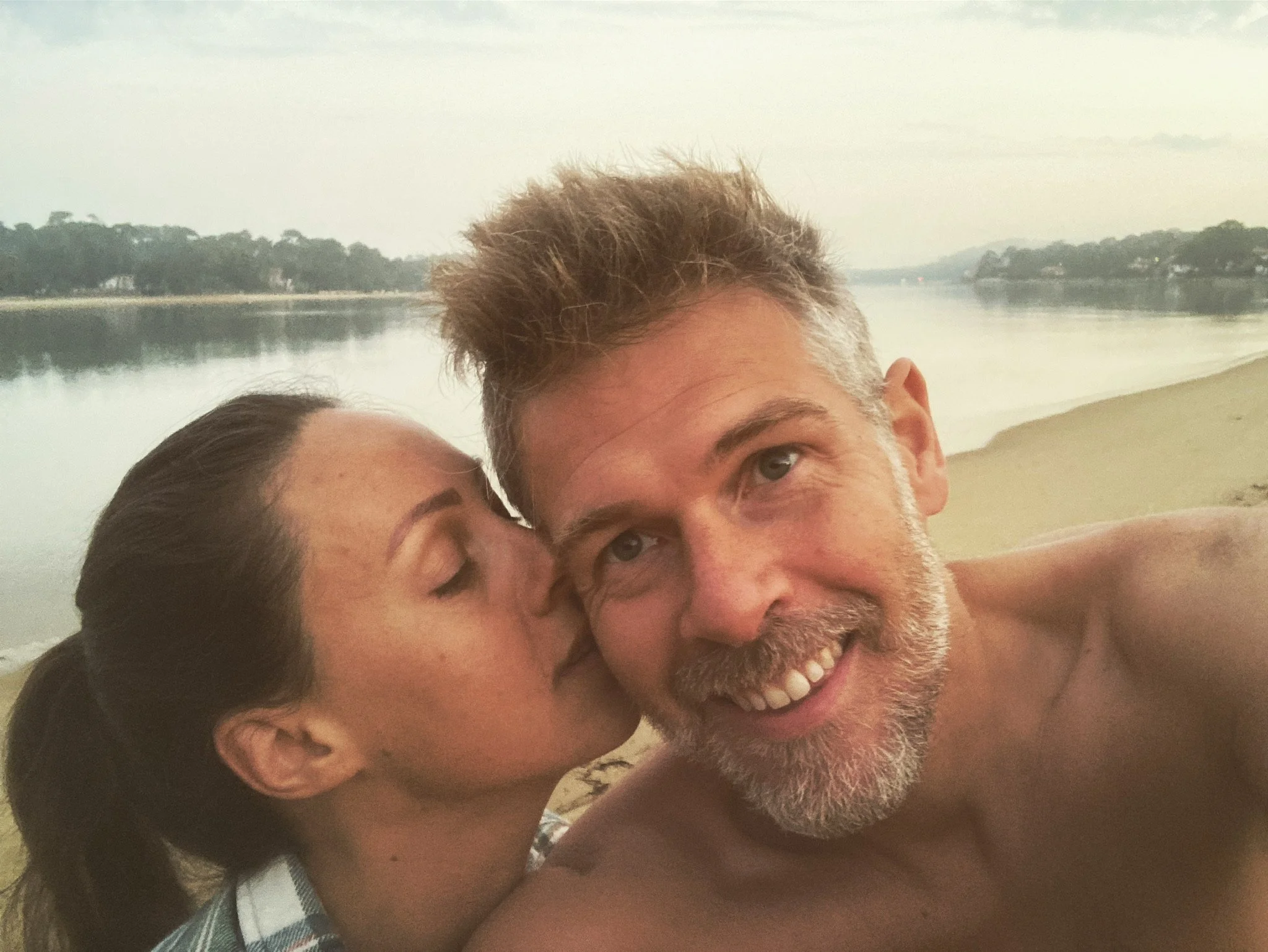 A woman kisses a shirtless man on the cheek while taking a selfie on a beach near a body of water with trees in the background.