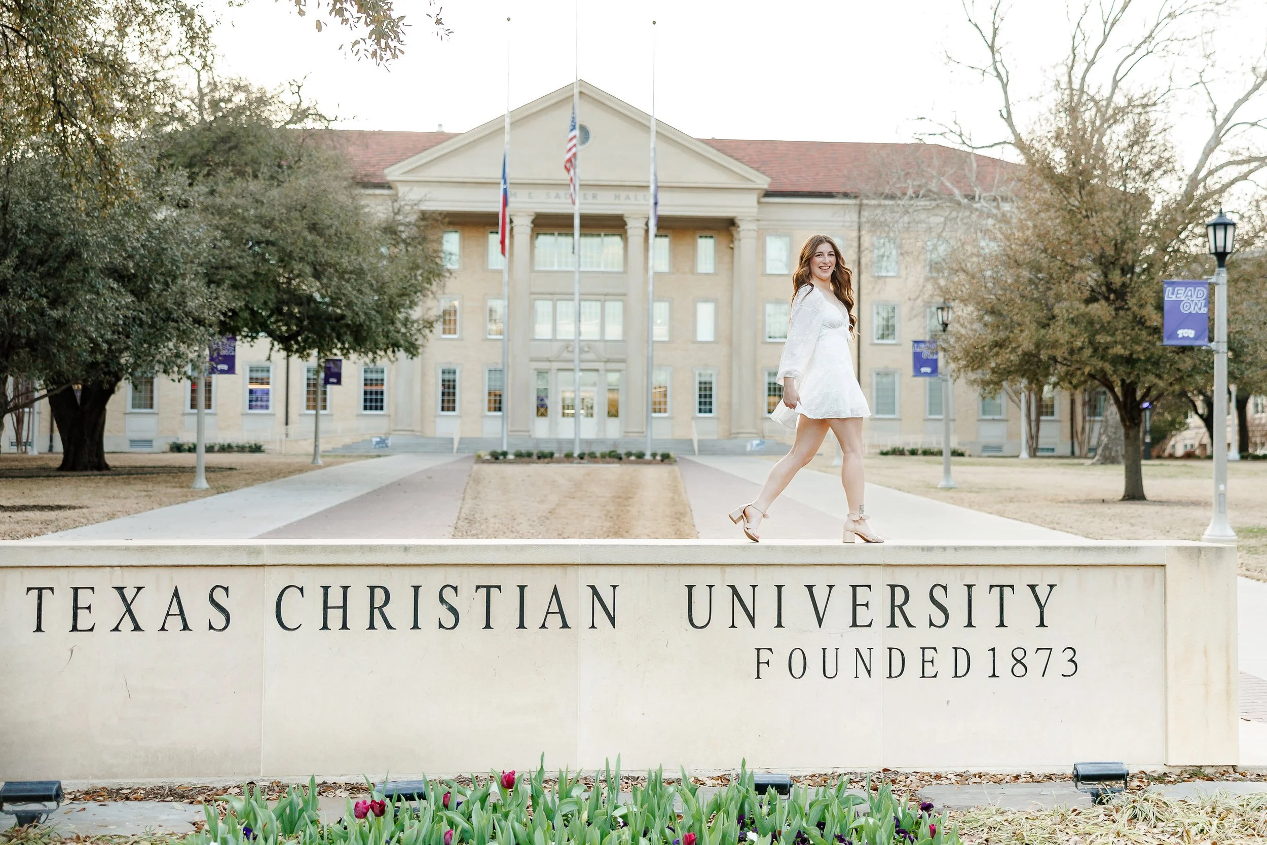 A smiling graduate in a white dress walks confidently across the iconic Texas Christian University sign in front of Sadler Hall. This joyful moment captures classic grad photos at Texas Christian University