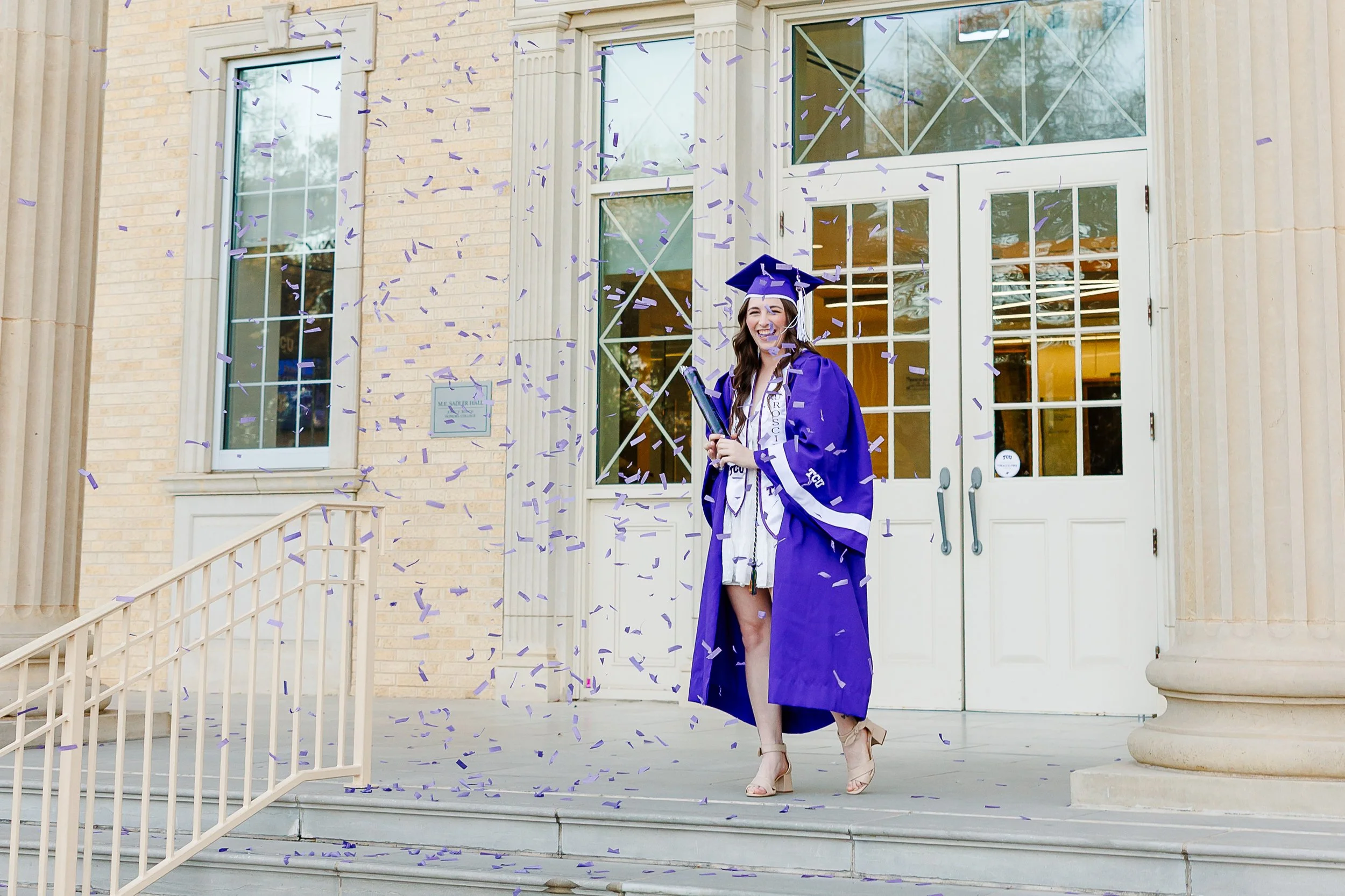 A joyful graduate in a purple cap and gown stands on the steps of a Texas Christian University building as purple confetti rains down around her, capturing a celebratory moment during grad photos at Texas Christian University.