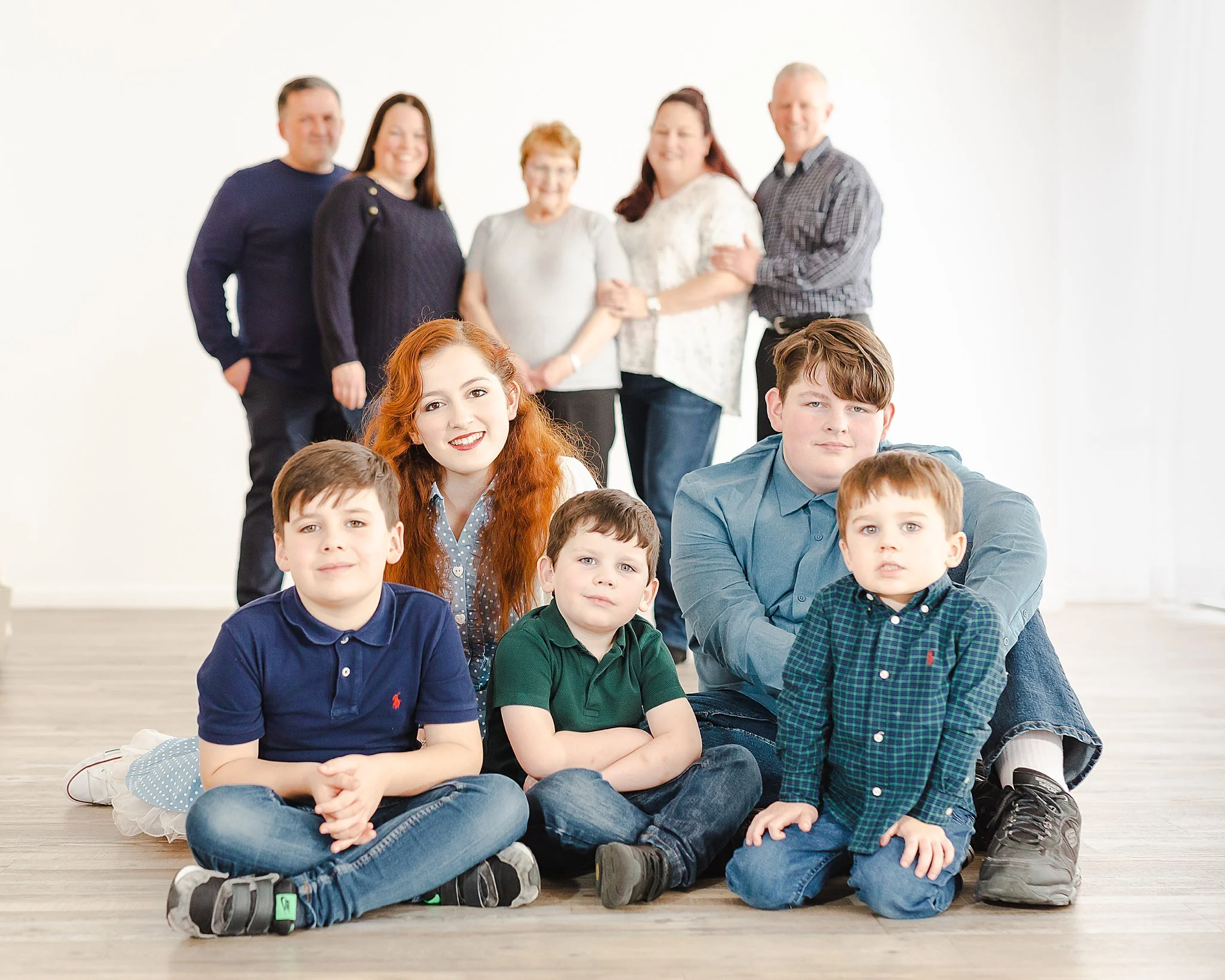 extended family portrait session of a family of ten with parents and grandparents blurred in the background and 5 grandkids in focus in the foreground. Parents are smiling down at the kids who are smiling at the camera.