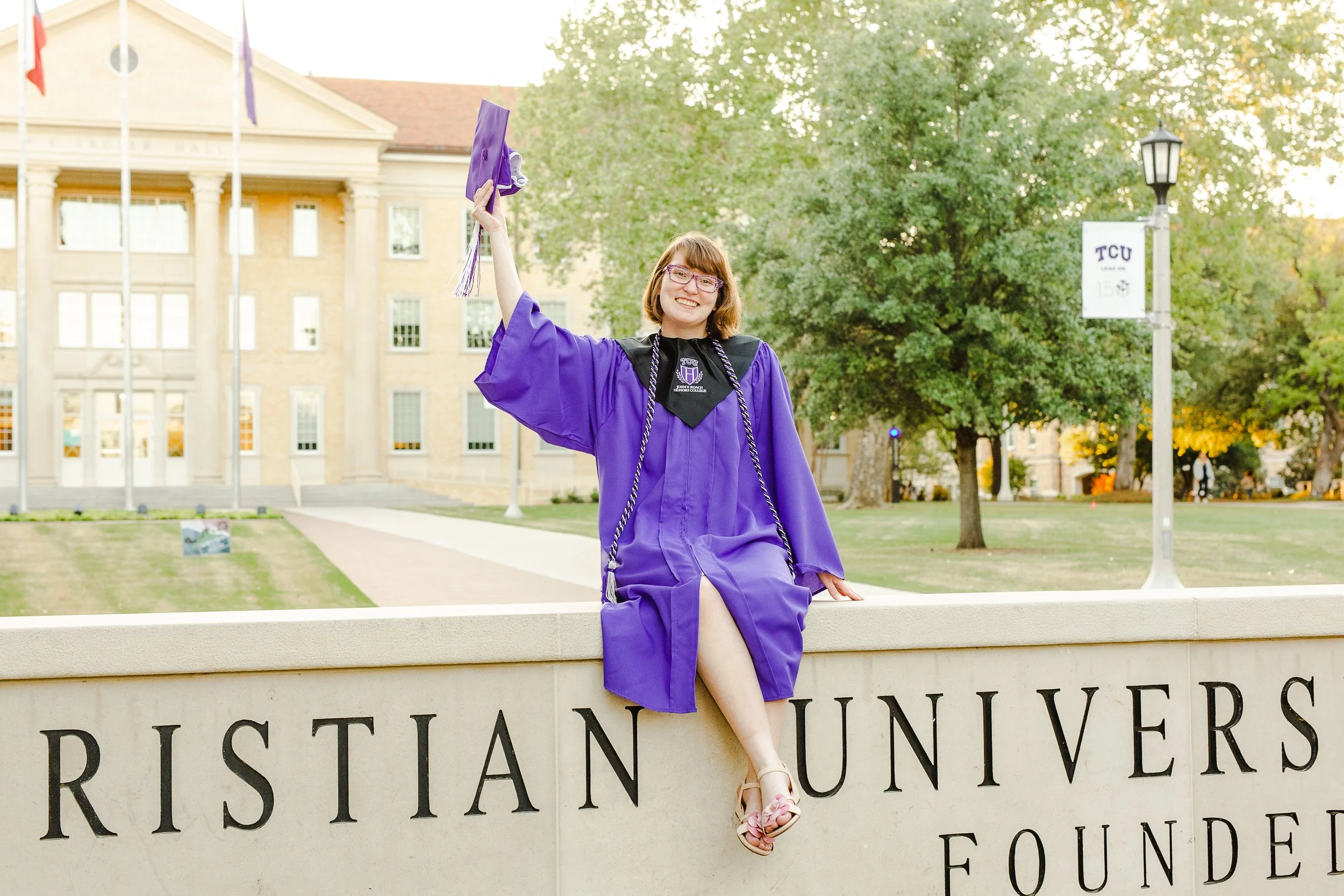Graduate in a purple gown and honor cords sits atop the Texas Christian University sign, holding her cap in the air and smiling proudly in front of Sadler Hall.