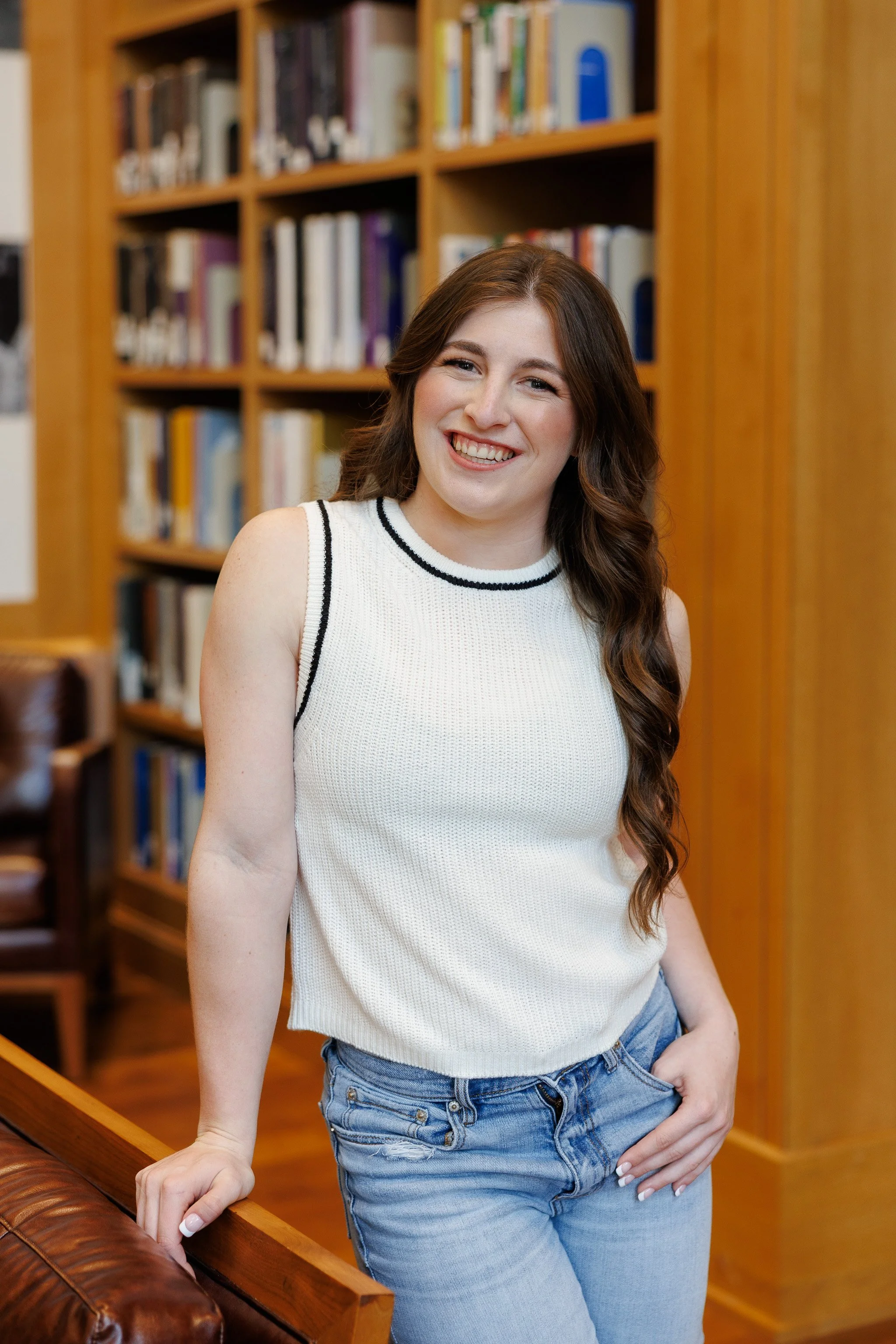 senior girl in a white sleeveless sweater and jeans standing in front of books inside the TCU Library posing for senior pictures