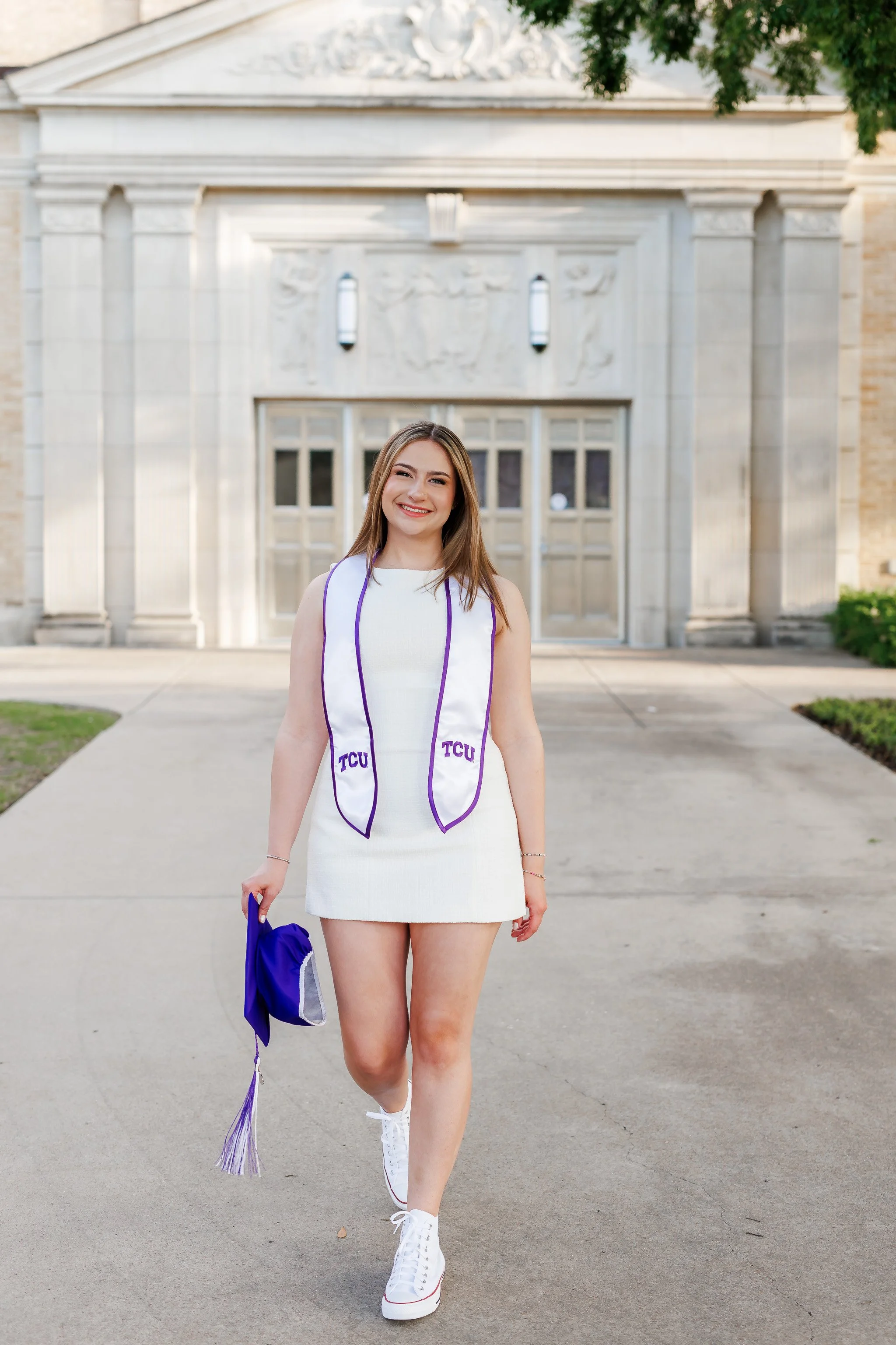 Senior girl wearing white dress walking with a graduation cap in front of Ed Landreth Hall at TCU posing for senior pictures
