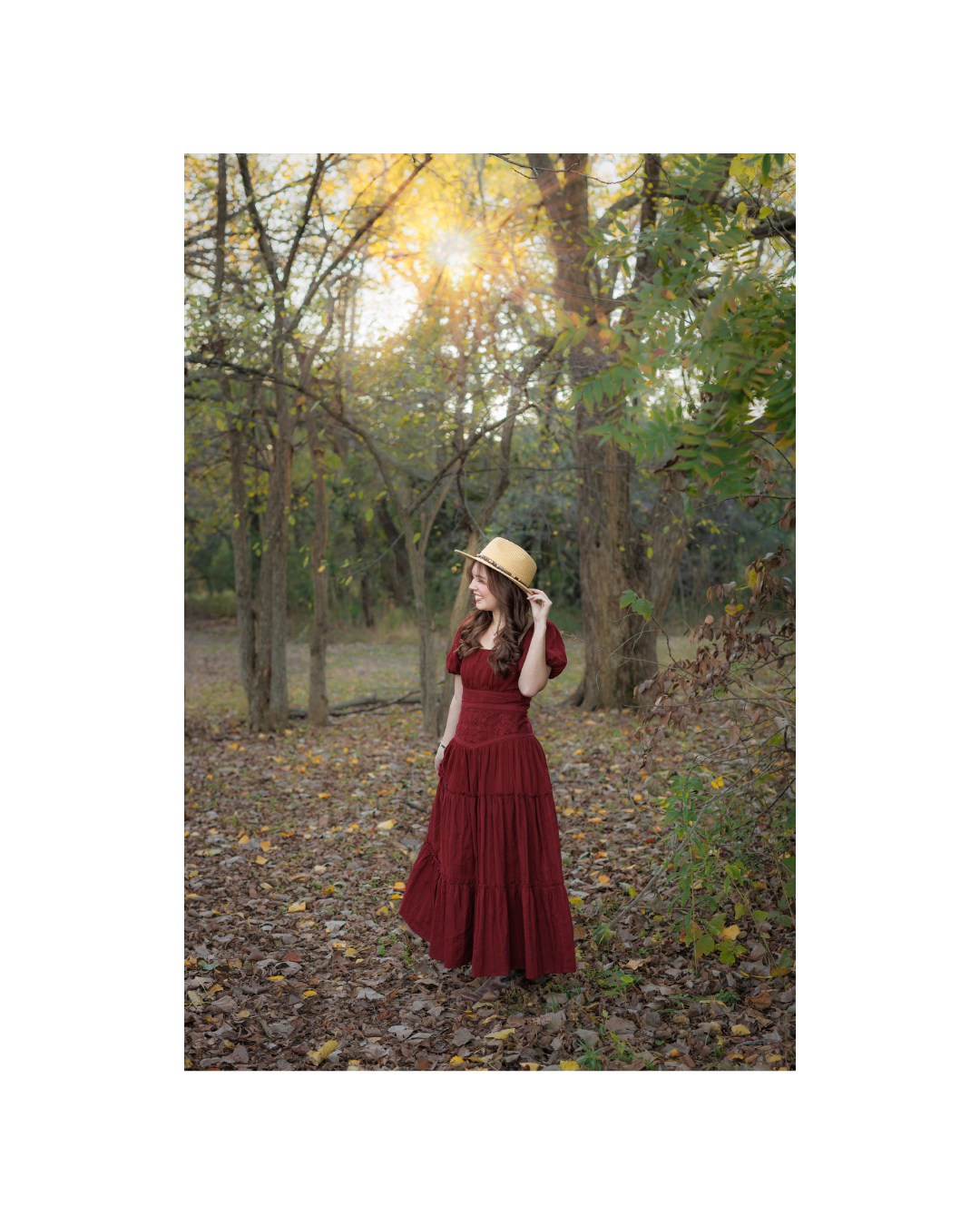 A young woman in a long red dress and a straw hat standing in a wooded area with fallen leaves, sunlight filtering through trees posing for senior portraits at Colleyville Nature Center by DeAndra Jarboe Photography.