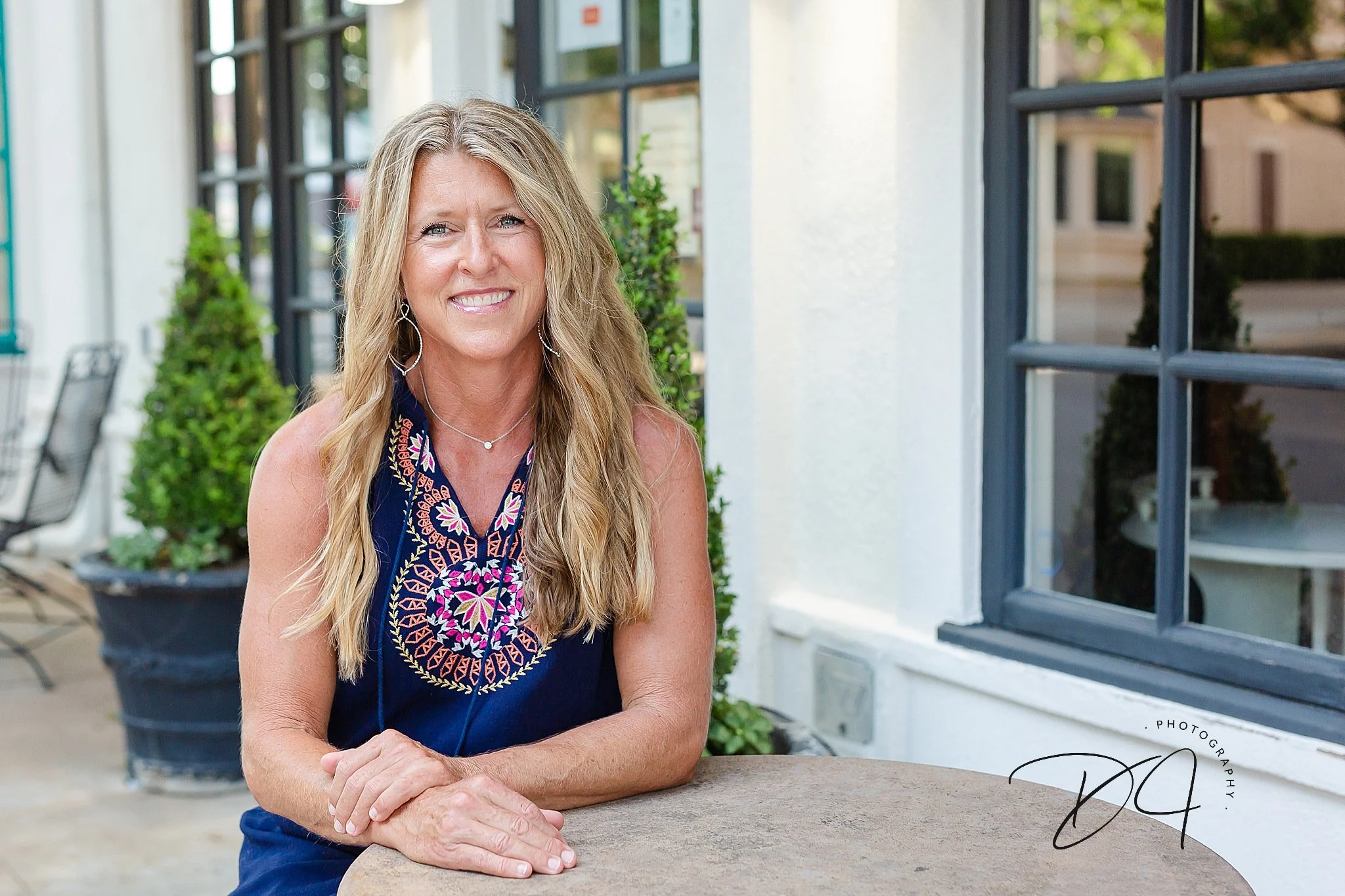 adult woman with long blond hair with beach waves wearing a dark blue sleeveless dress and statement earrings sitting at a bistro table on a sidewalk posing for headshots at Colleyville Library