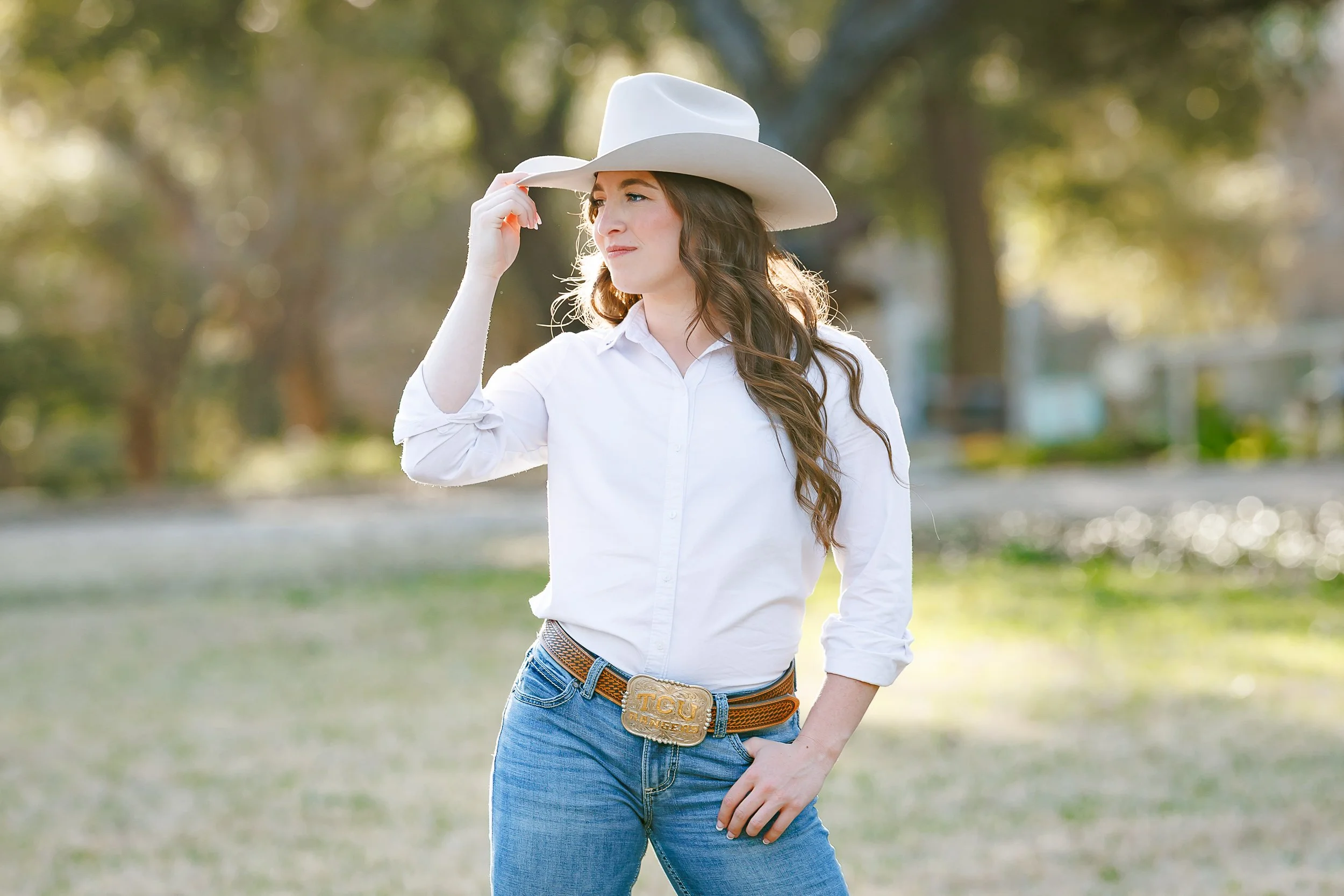 A graduate in a cowboy hat and TCU-branded belt buckle gently tips her hat while standing in the golden light on campus. A bold and stylish representation of grad photos at Texas Christian University