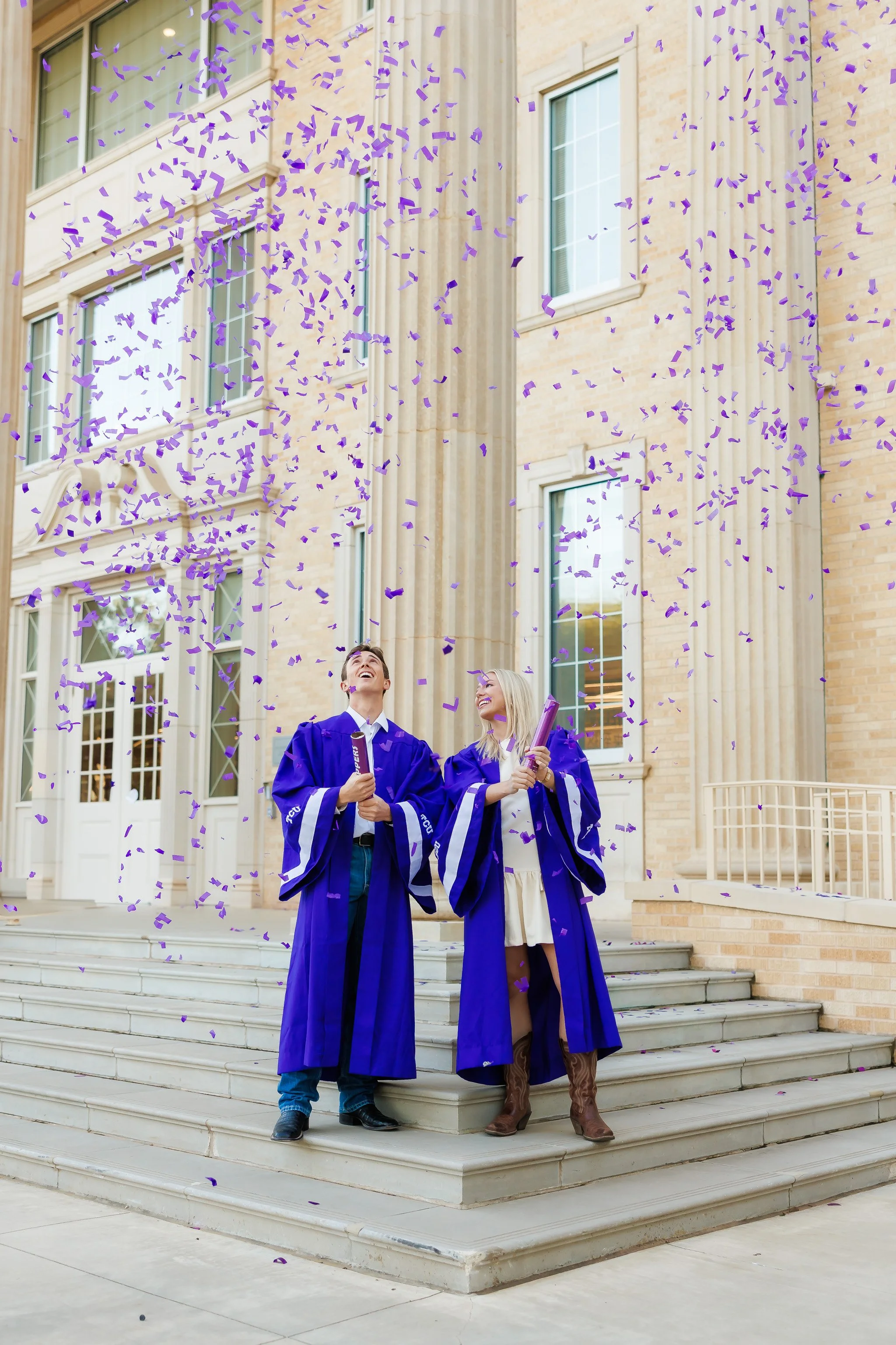 TCU senior guy and senior girl wearing purple graduation gowns and popping confetti on the steps of Sadler Hall for senior pictures