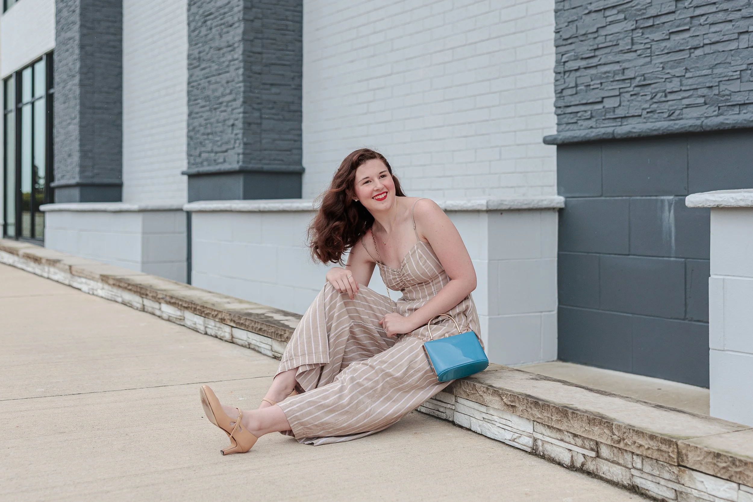 Young woman in a striped jumpsuit sitting on a low ledge outside a modern building, smiling with a relaxed posture and holding a blue purse, capturing graduation photos while choosing to pose naturally.