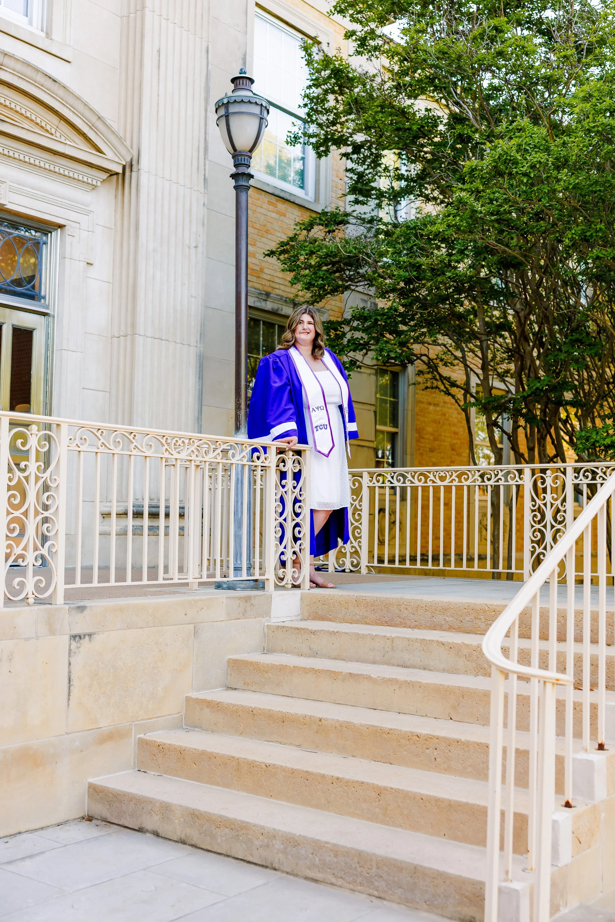 TCU senior girl in white dress and purple graduation gown standing on steps at the entrance to the Ed Landreth Hall for graduation portraits