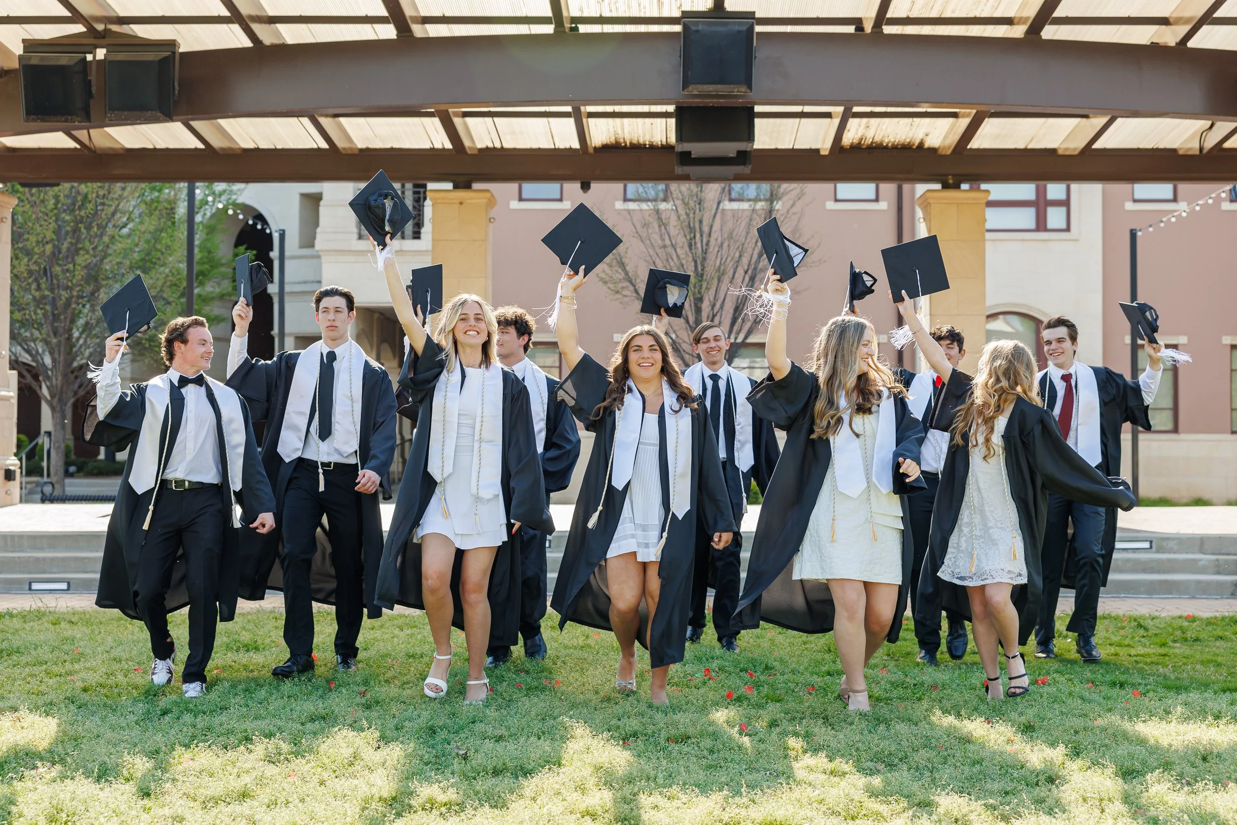 A group of ten young people, four women and six men, dressed in graduation gowns, walking towards the camera holding caps in the air cheerfully on a sunny day at the Colleyville Library. They are posing for senior class pictures.