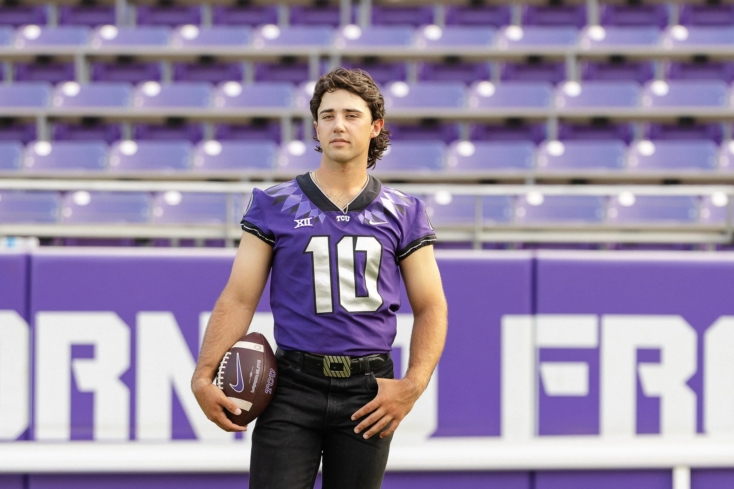 Young man in TCU football jersey holding a football while standing confidently in the stadium, capturing pride and accomplishment in TCU football graduation photos; photographed by Dallas photographer.