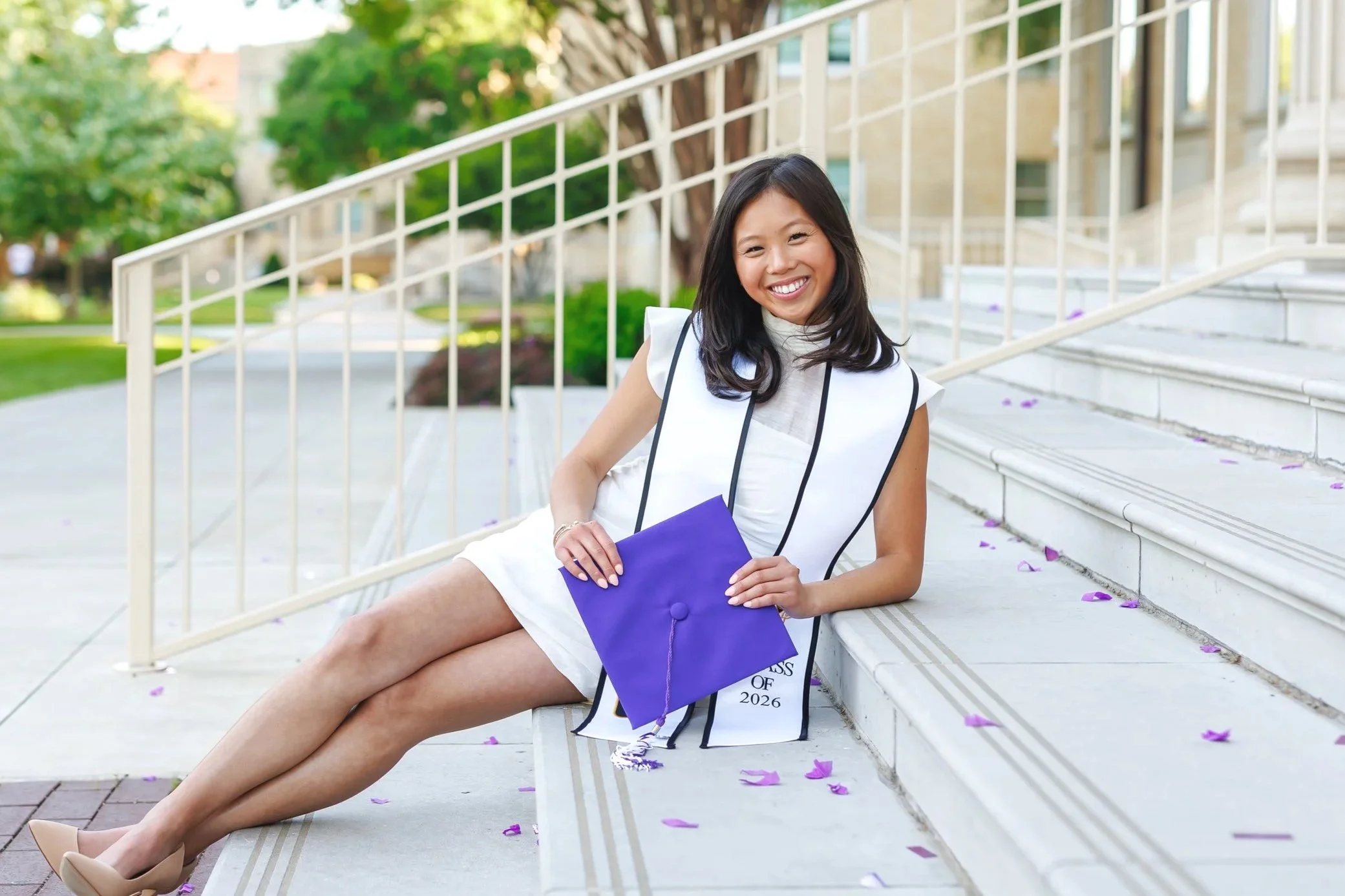 Senior girl at TCU wearing short white dress lounging on the steps in front of Sadler Hall posing for senior pictures at Texas Christian University smiling at the camera while holding a purple horn front graduation cap.