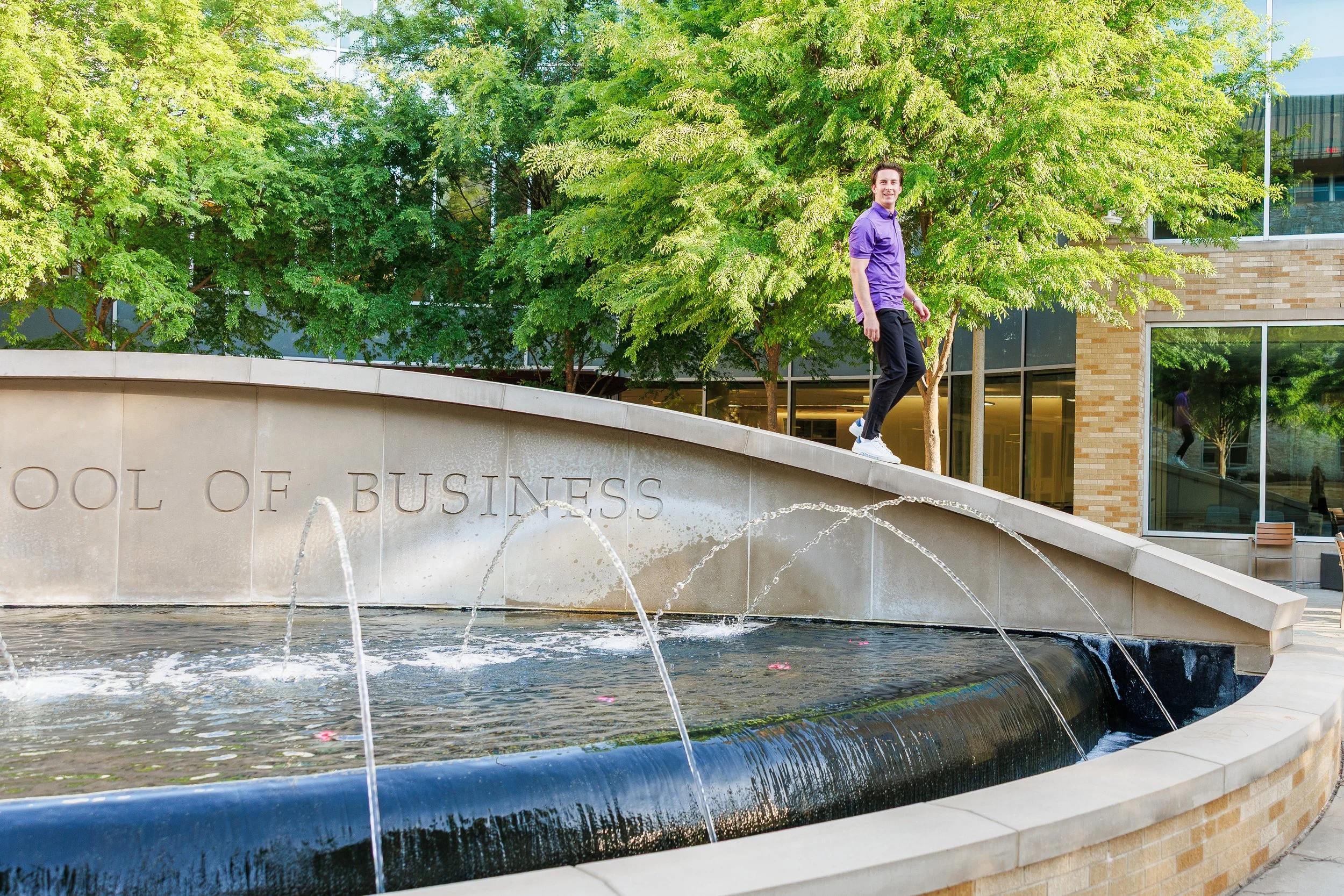 TCU senior guy standing atop the water fountain at Neeley School of Business for Fort Worth graduation portraits