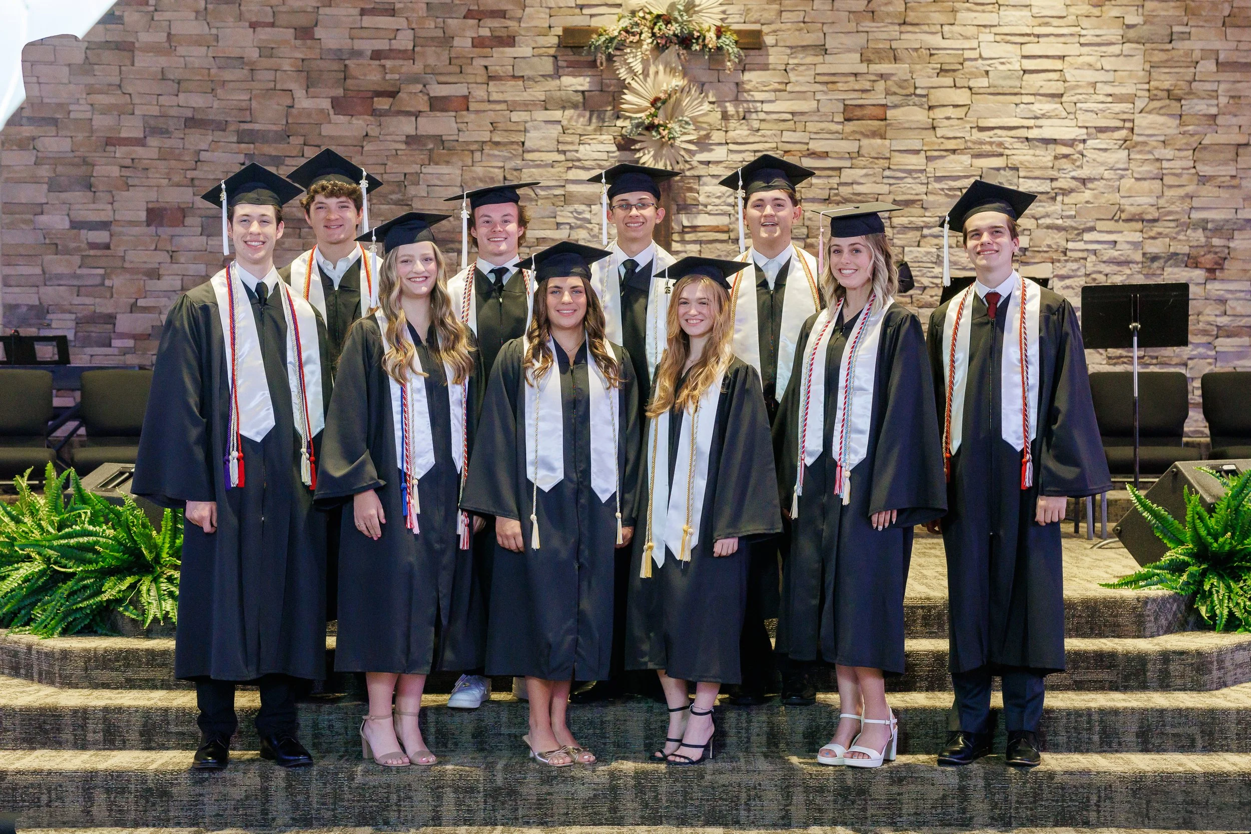 group of ten highschool seniors on graduation day posing formally for a class pictures wearing black graduation gowns, white stoles, gold honor cords and mortar boards. Everyone is smiling at the camera standing inside a church in Bedford, Texas.