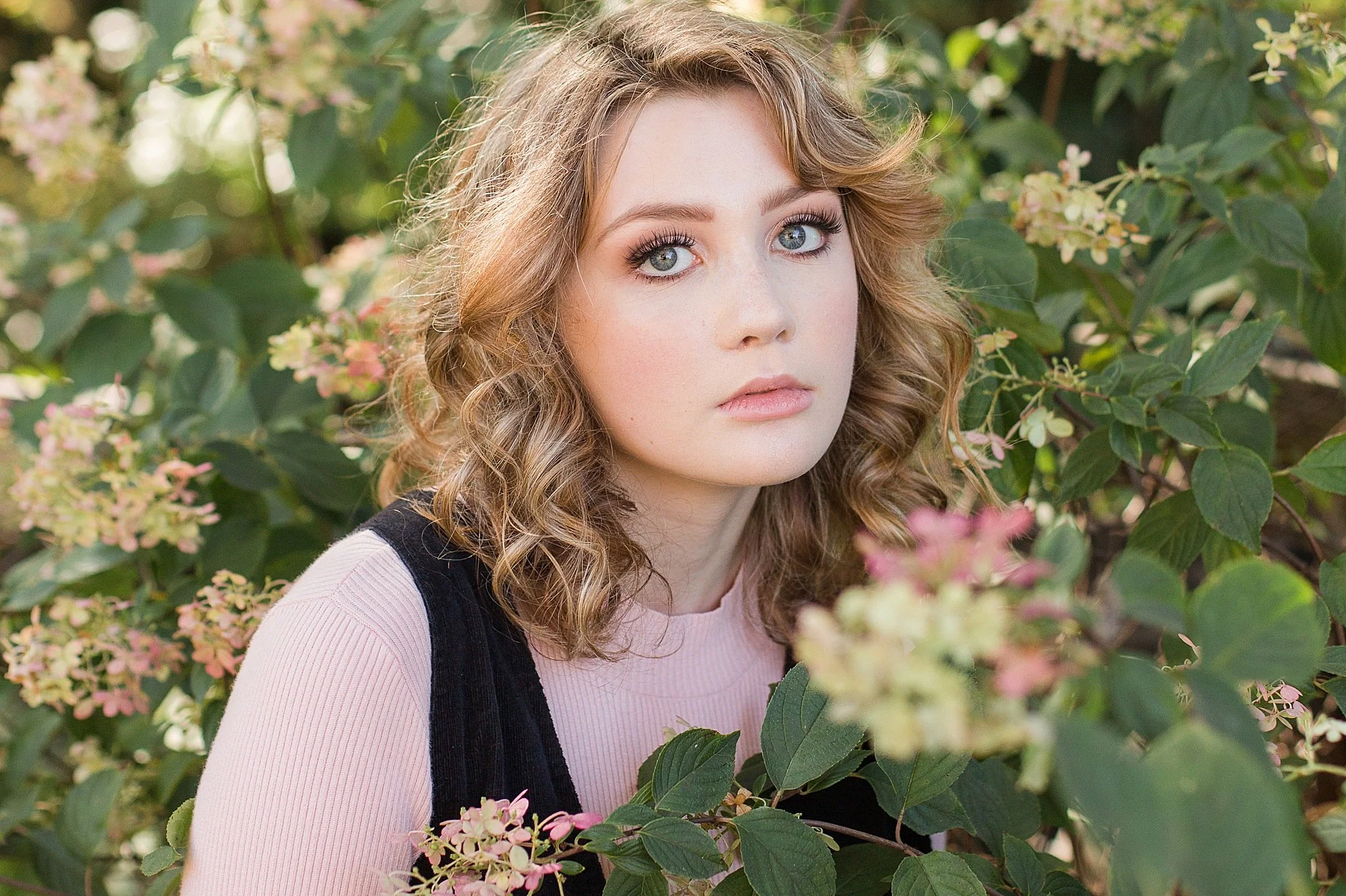 young lading with light brown wavy hair and big green eyes wearing a pink top and black jumpsuit staring somberly at the camera tucked in a pink hydrangea bush posing for senior pictures with DeAndra Jarboe Photography