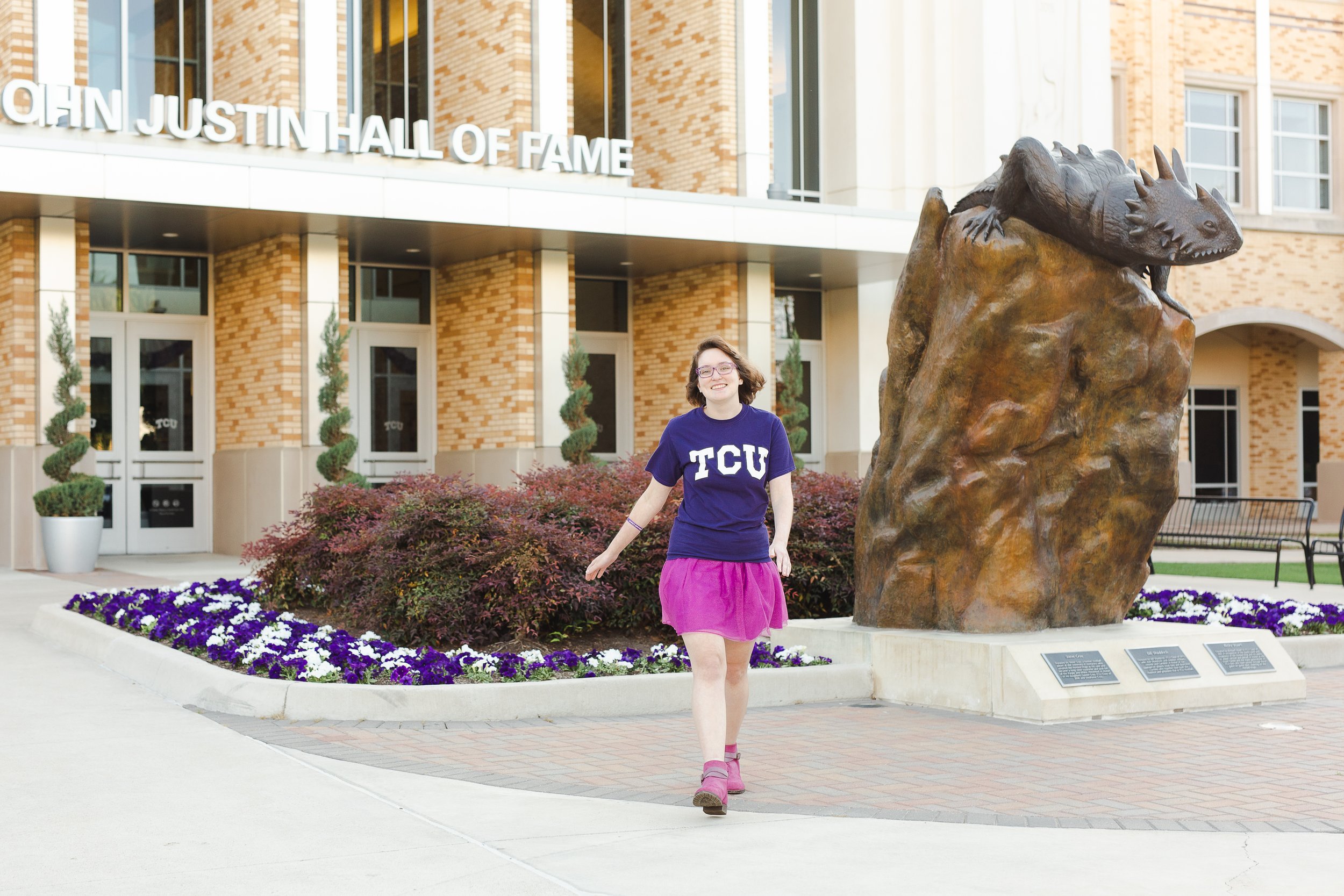 Student wearing a TCU shirt and pink skirt walks confidently in front of the Horned Frog statue and John Justin Hall of Fame, demonstrating relaxed and natural graduation photo pose ideas.