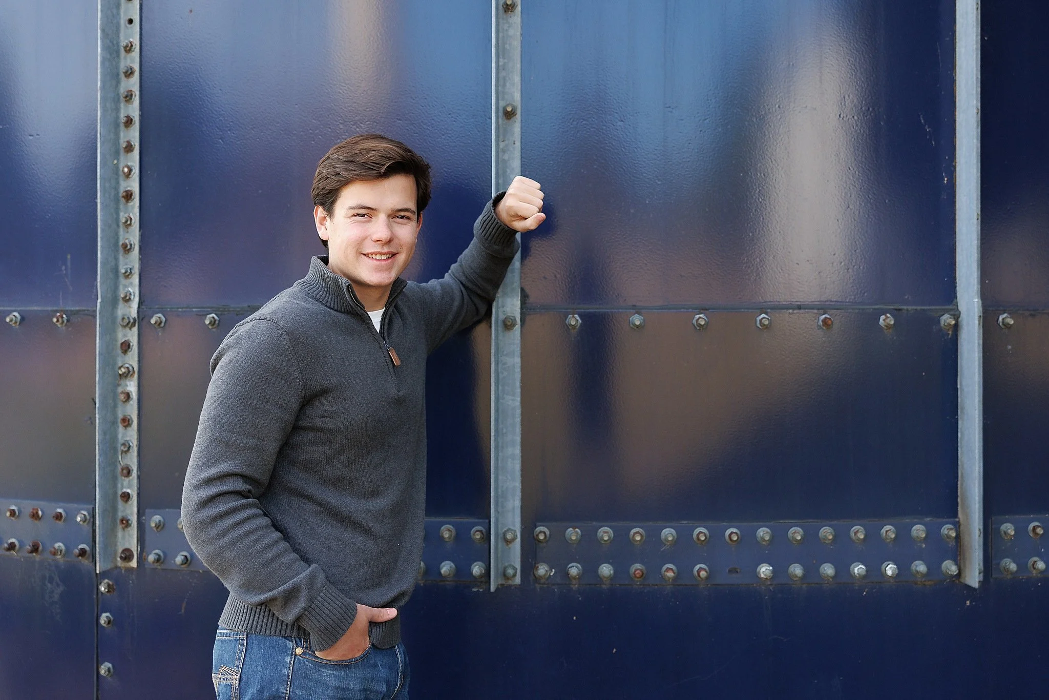 young man with dark short hair in a gray quarter zip top and jeans leaning against a navy blue metal building posing for senior pictures at McPherson Park