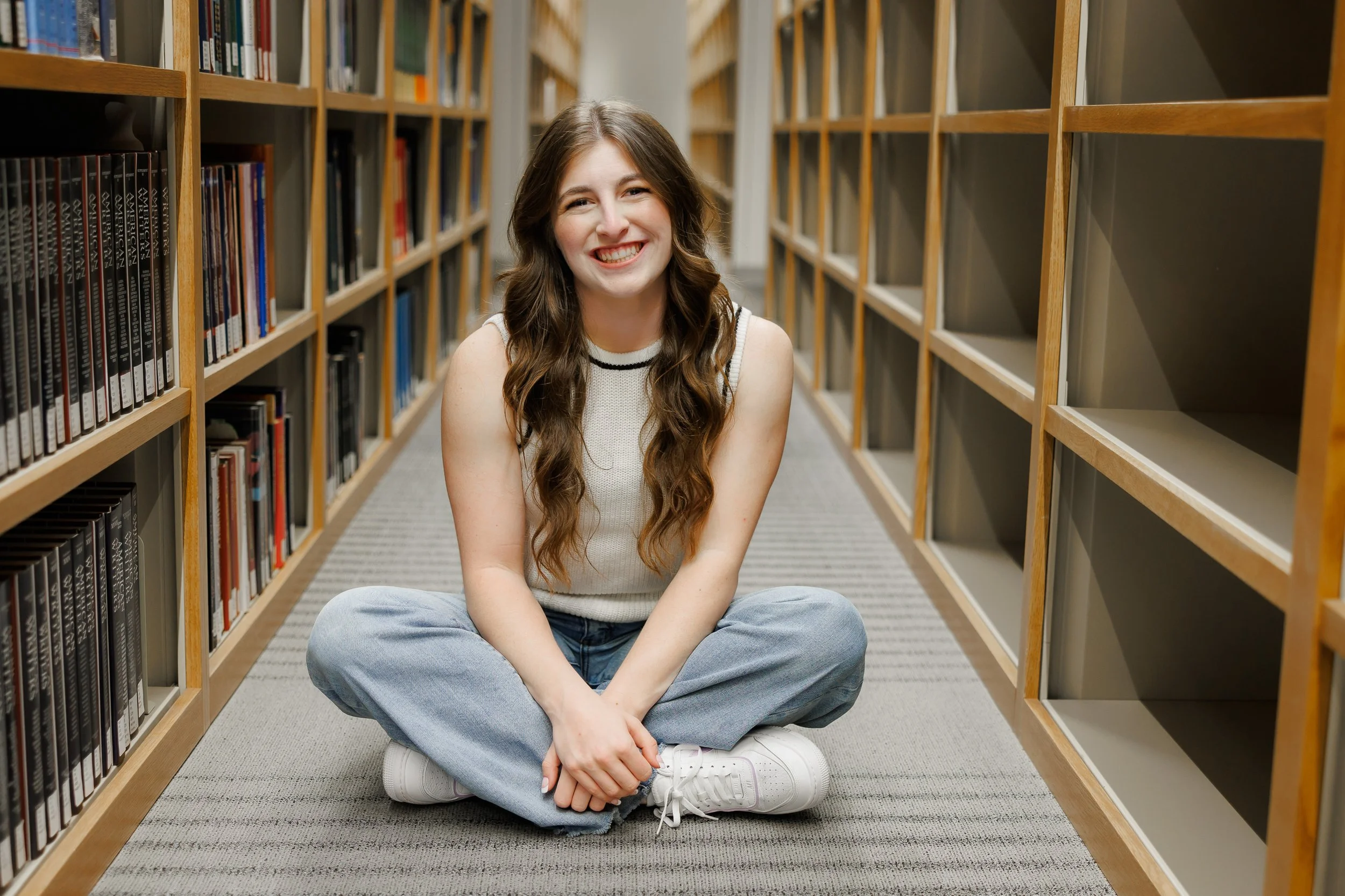 Senior girl in a white sleeveless sweater and jeans sitting between rows of books inside the TCU Library posing for senior pictures