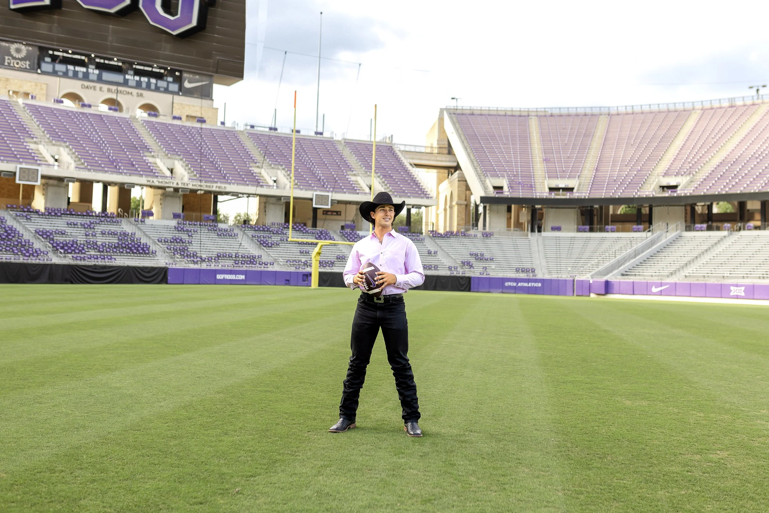 Wide shot of graduate standing confidently at midfield in an empty TCU stadium, wearing a pink shirt and black cowboy hat while holding a football