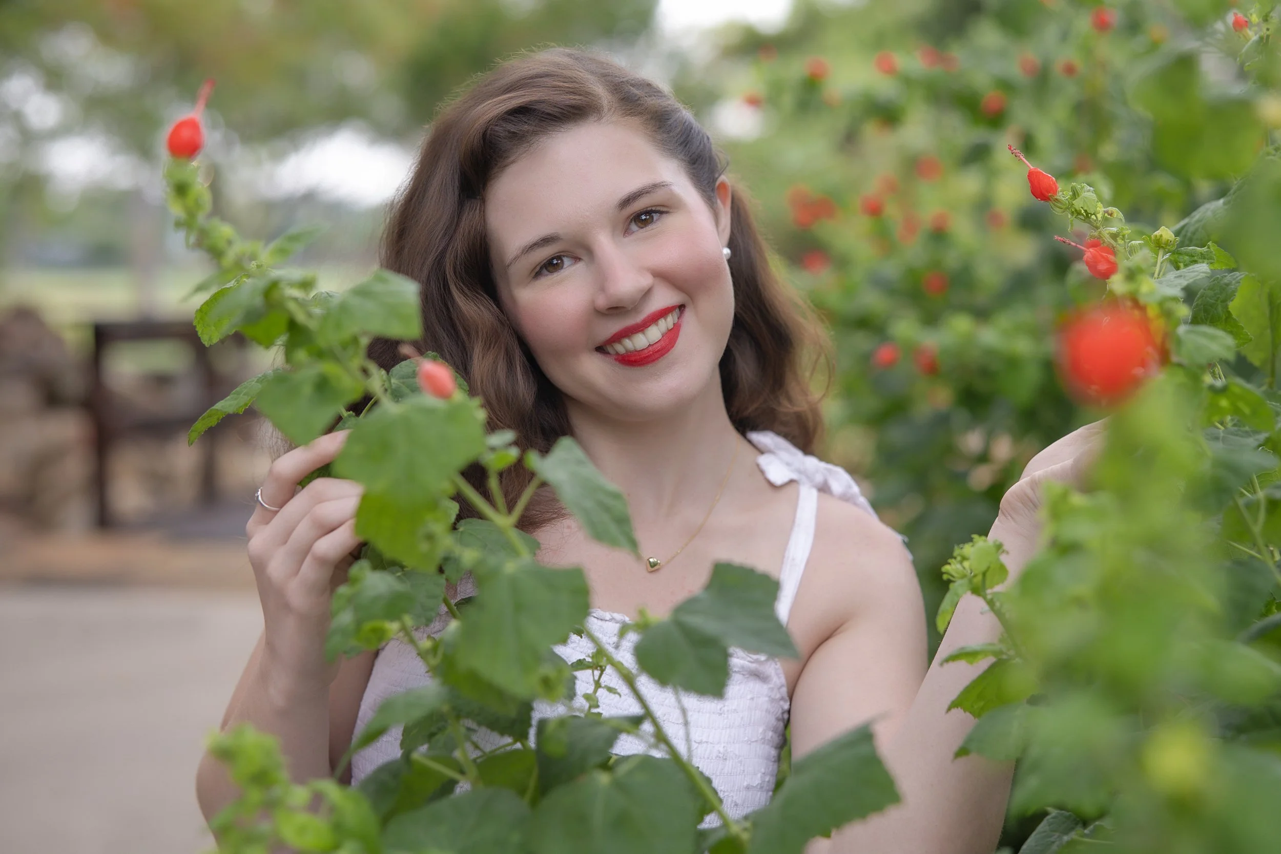 Young woman smiling in a garden surrounded by green foliage and red flower buds, wearing a white dress and gold necklace, showcasing graduation photos while posing naturally in an outdoor setting.